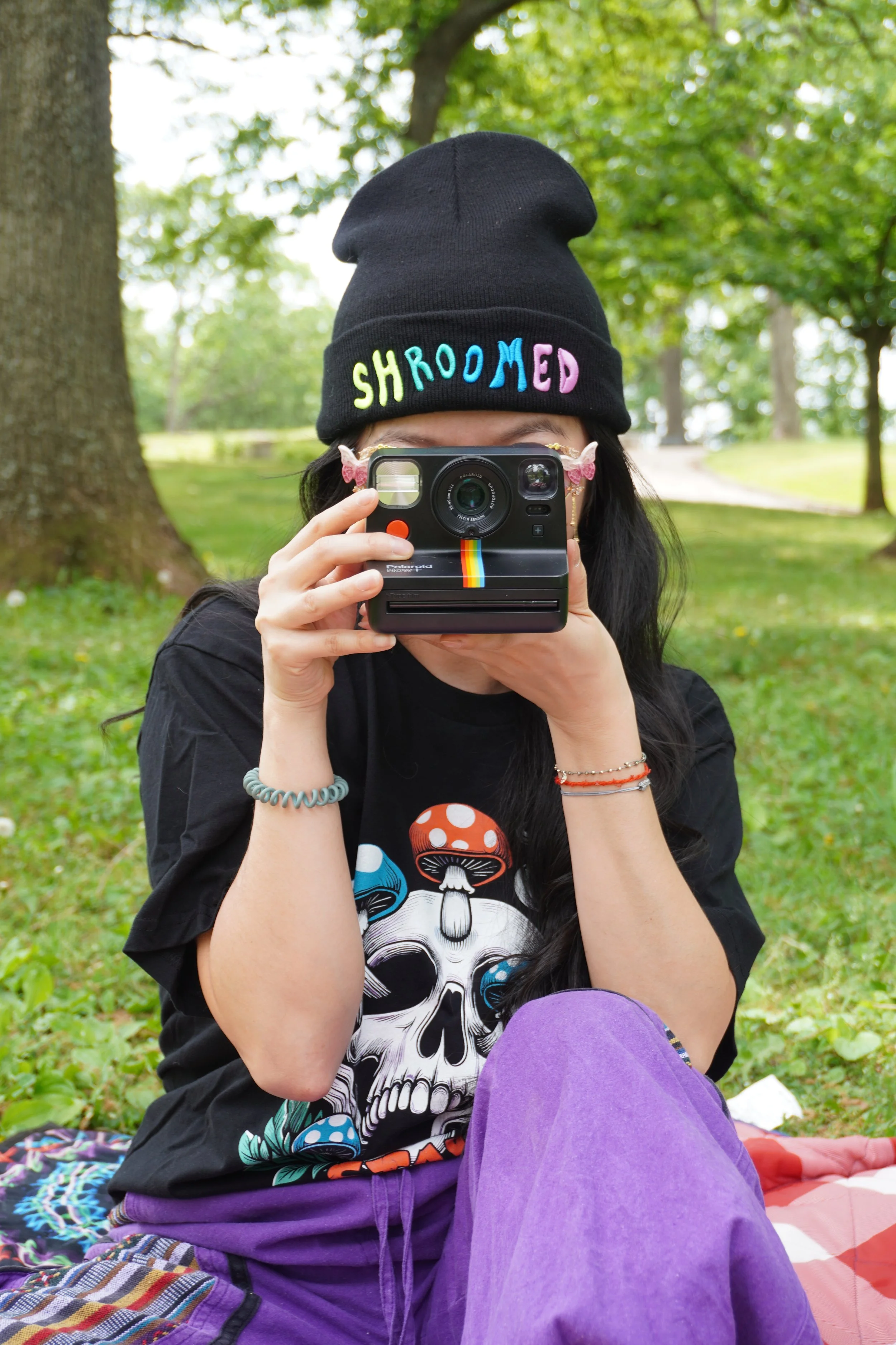 Person sitting outdoors in front of trees, wearing a black beanie with colorful embroidered letters spelling 'SHROOMED,' a skull graphic T-shirt, purple pants, holding a vintage instant film camera with a rainbow stripe, wearing butterfly earrings an