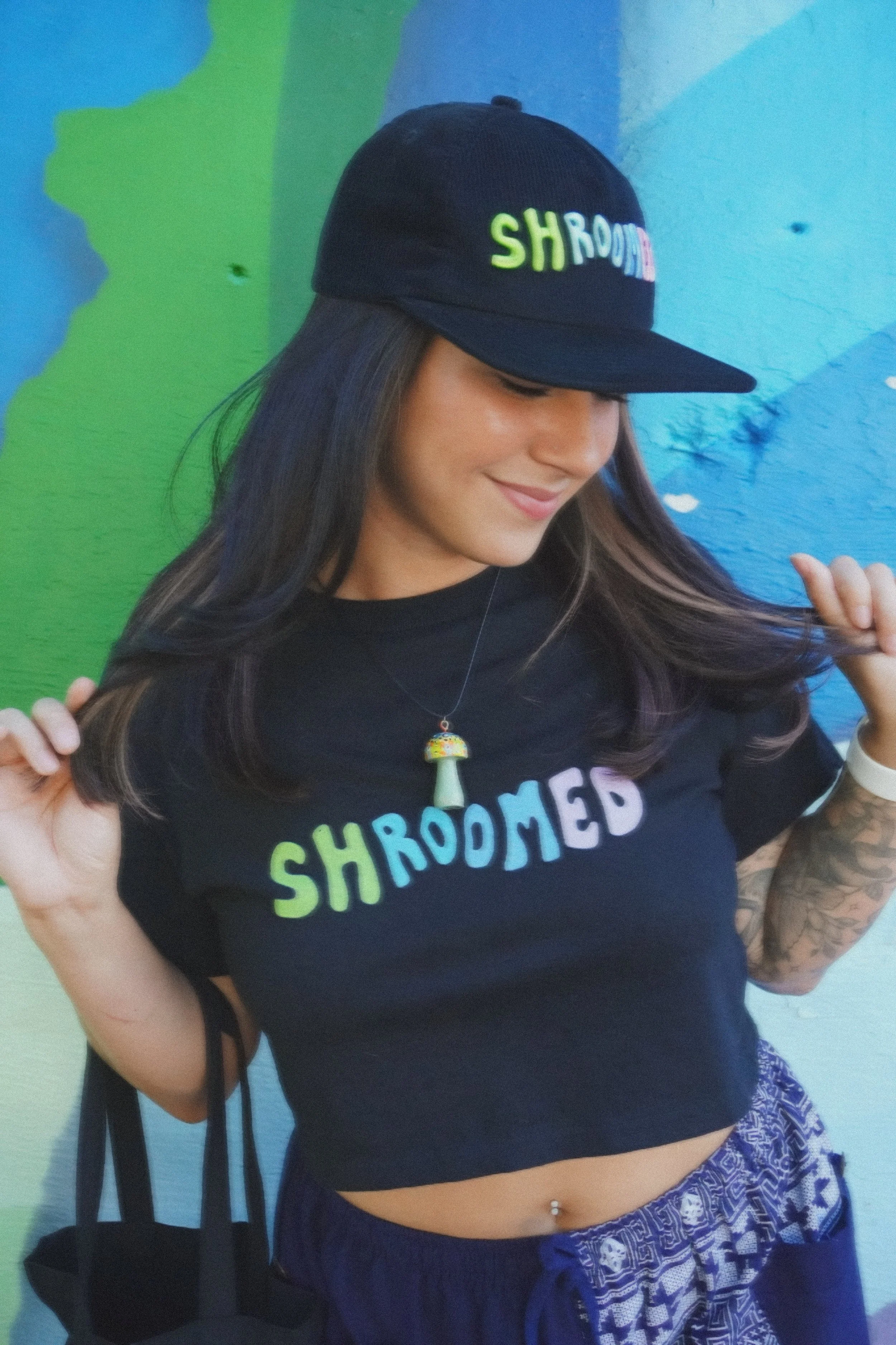 A young woman wearing a black baseball cap with the word 'SHROOOMED' embroidered on it, a black t-shirt with the same word, and a blue patterned skirt, standing in front of a colorful mural painted on a wall.
