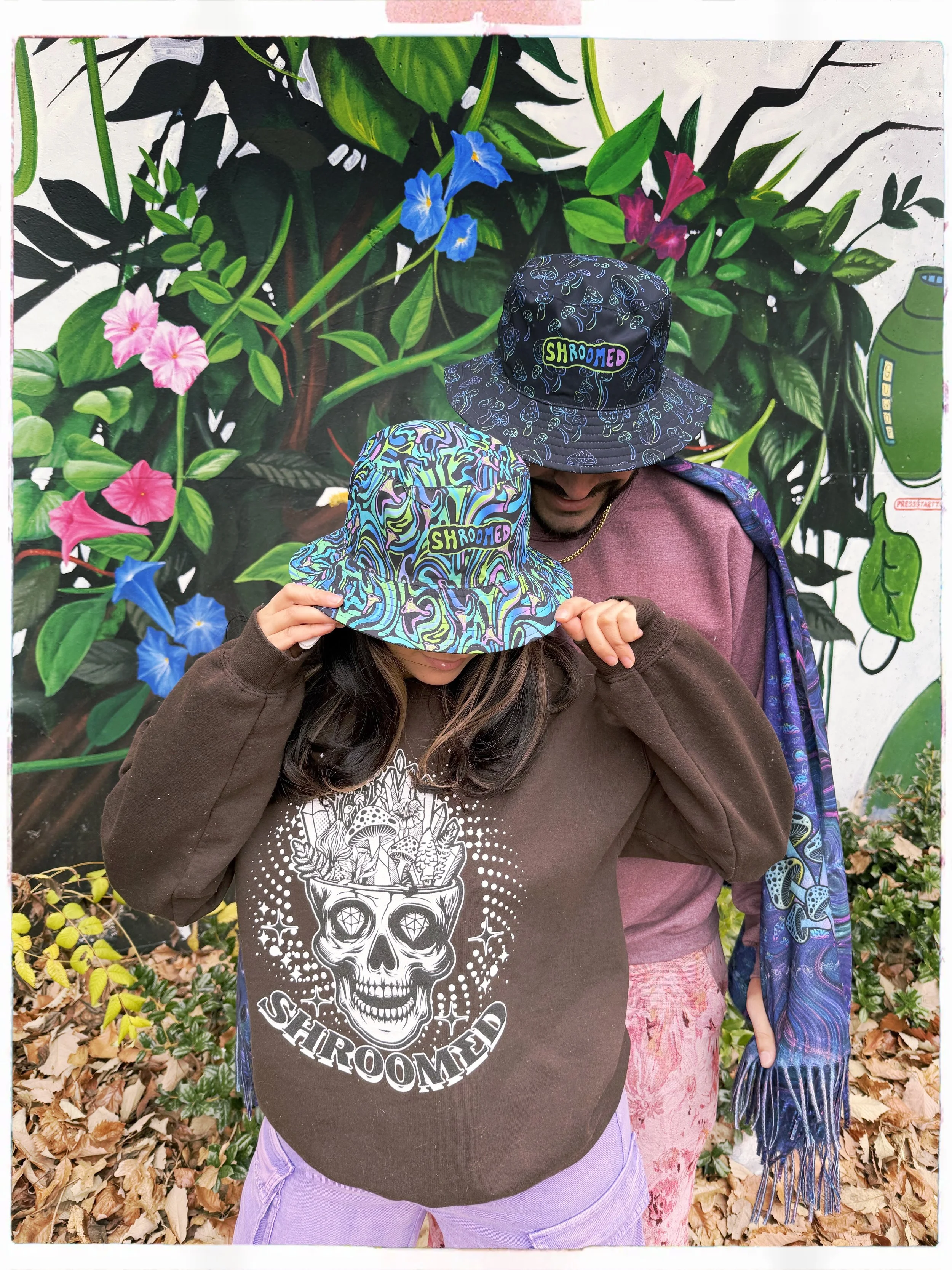 Two people wearing bucket hats with colorful designs and 'Shroomed' patches, standing outdoors in front of a mural of tropical plants and flowers.
