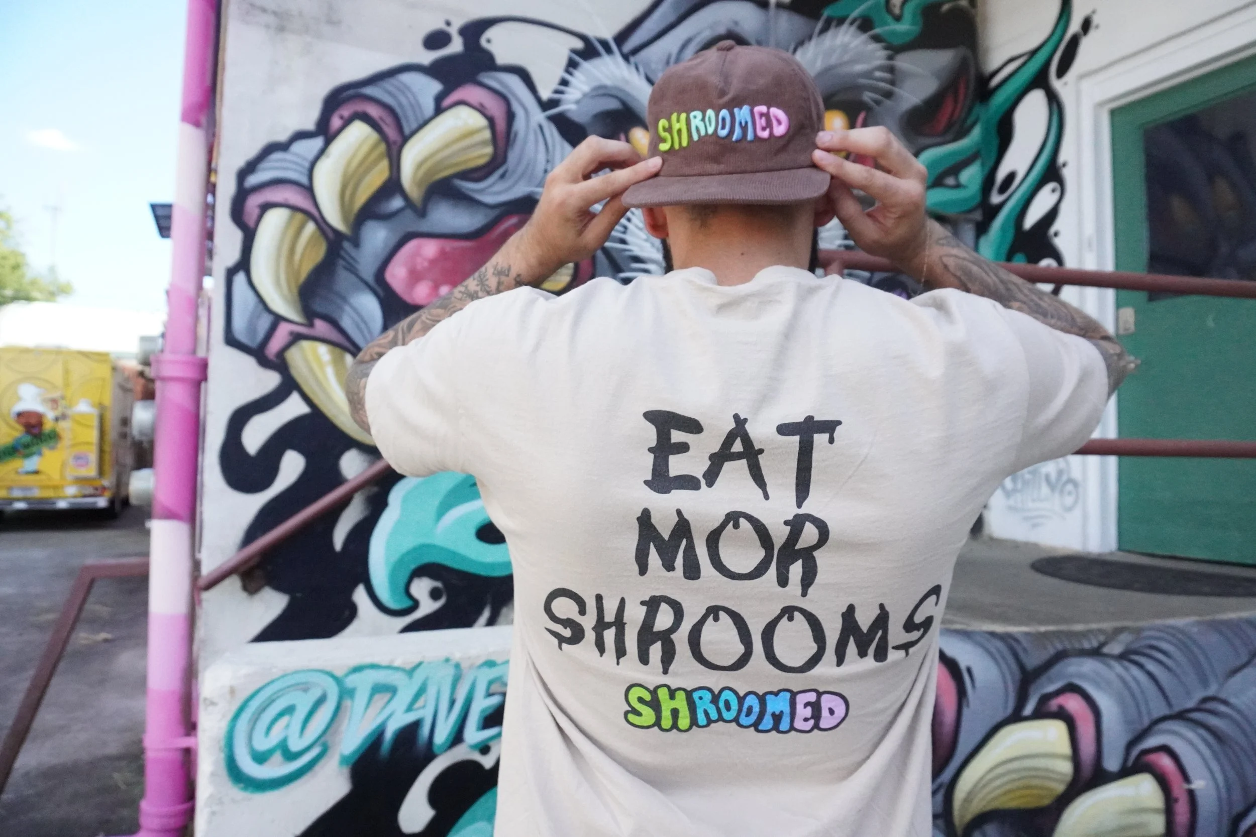 Person with tattoos adjusting a brown cap embroidered with colorful letters spelling 'SHROMMED' in front of a vibrant graffiti wall.