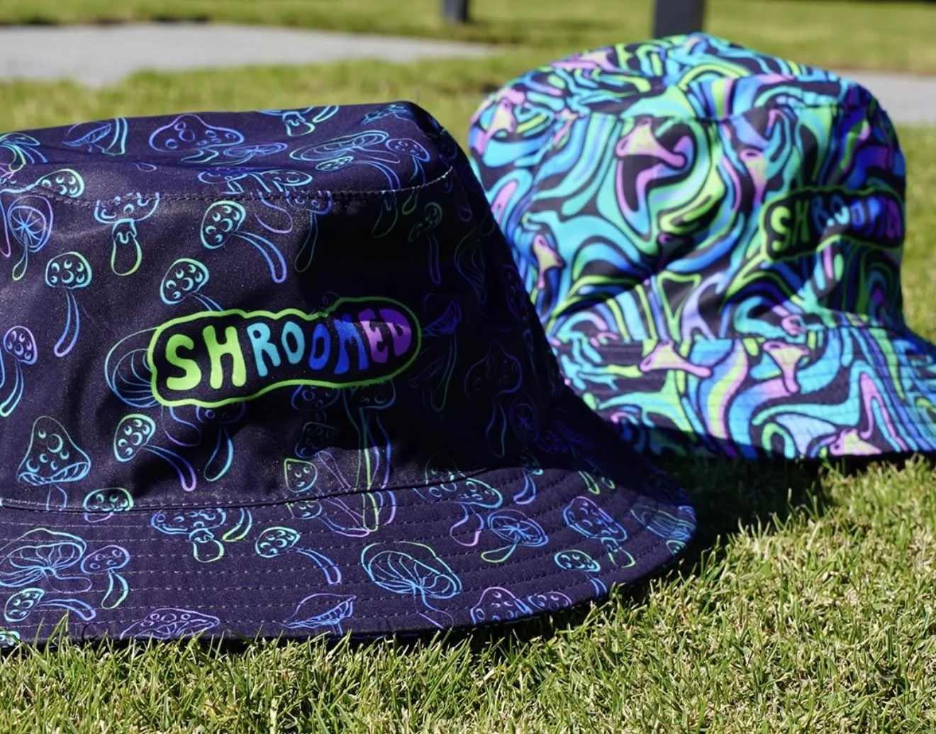 Two colorful bucket hats with neon mushroom and swirling designs, placed on grass outdoors.