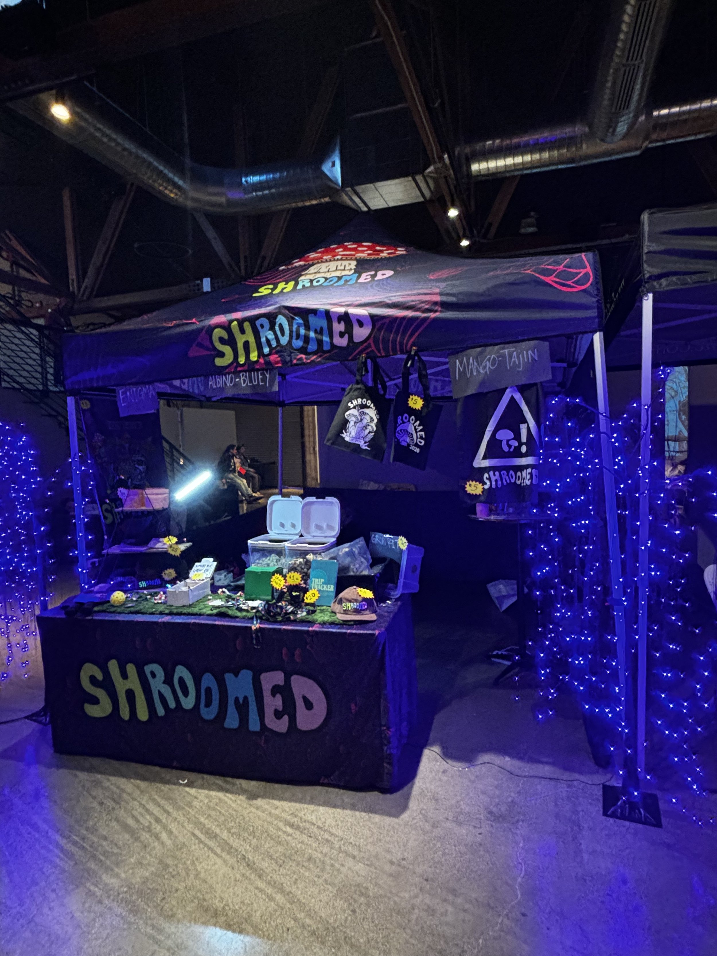 A booth decorated with black and purple lights, featuring purple string lights and a black tablecloth with colorful Shroomed lettering, set up at an indoor event.