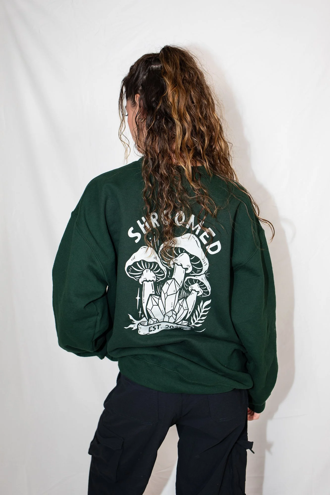 Person with long curly hair, wearing a black sweatshirt with graphic design of mushrooms, crystals, and the text 'Share the M' and 'Est. 2021', standing against a plain white background.