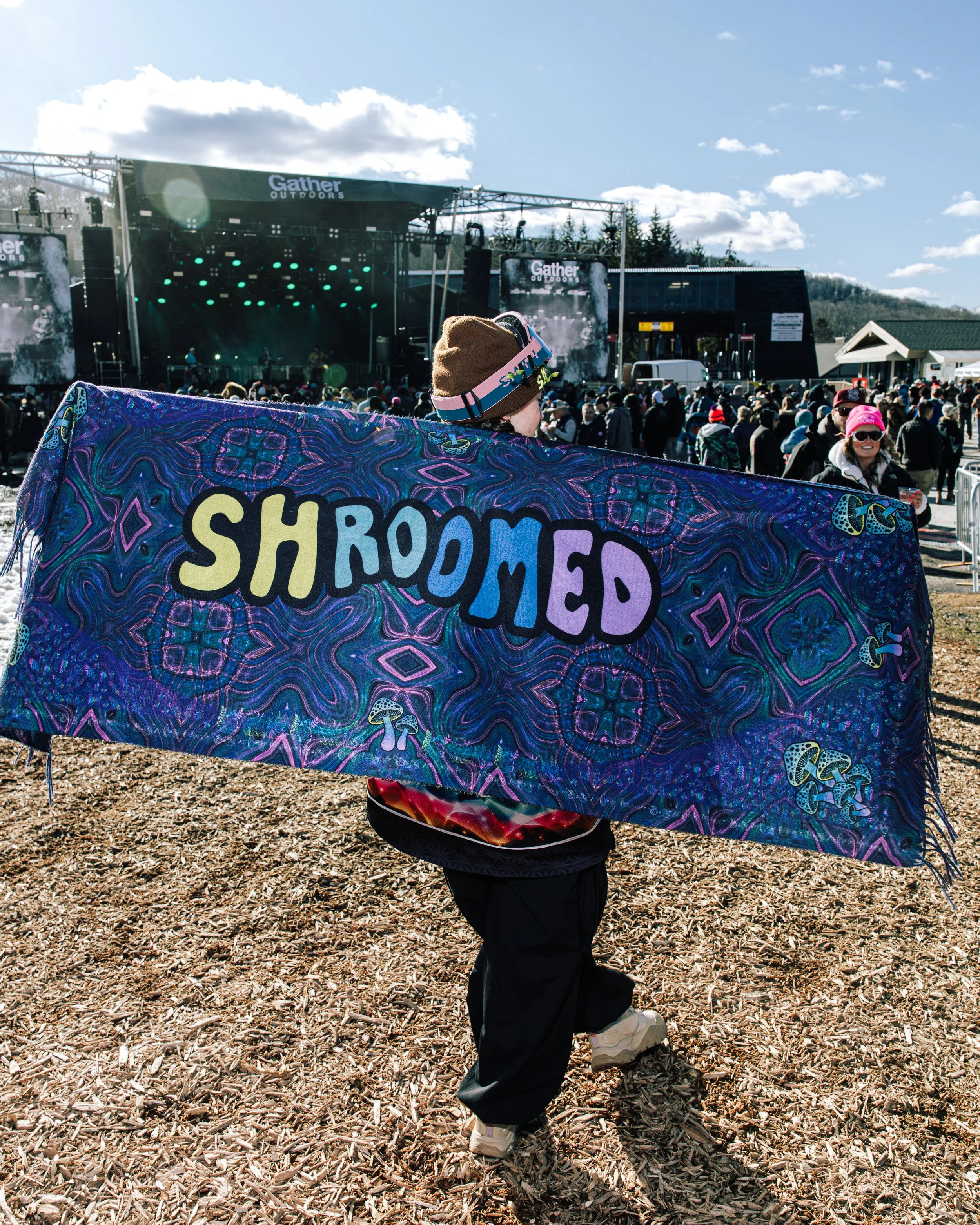 A person holding a colorful towel that reads "SHROOMED" at an outdoor concert stage with a crowd and a blue sky in the background.