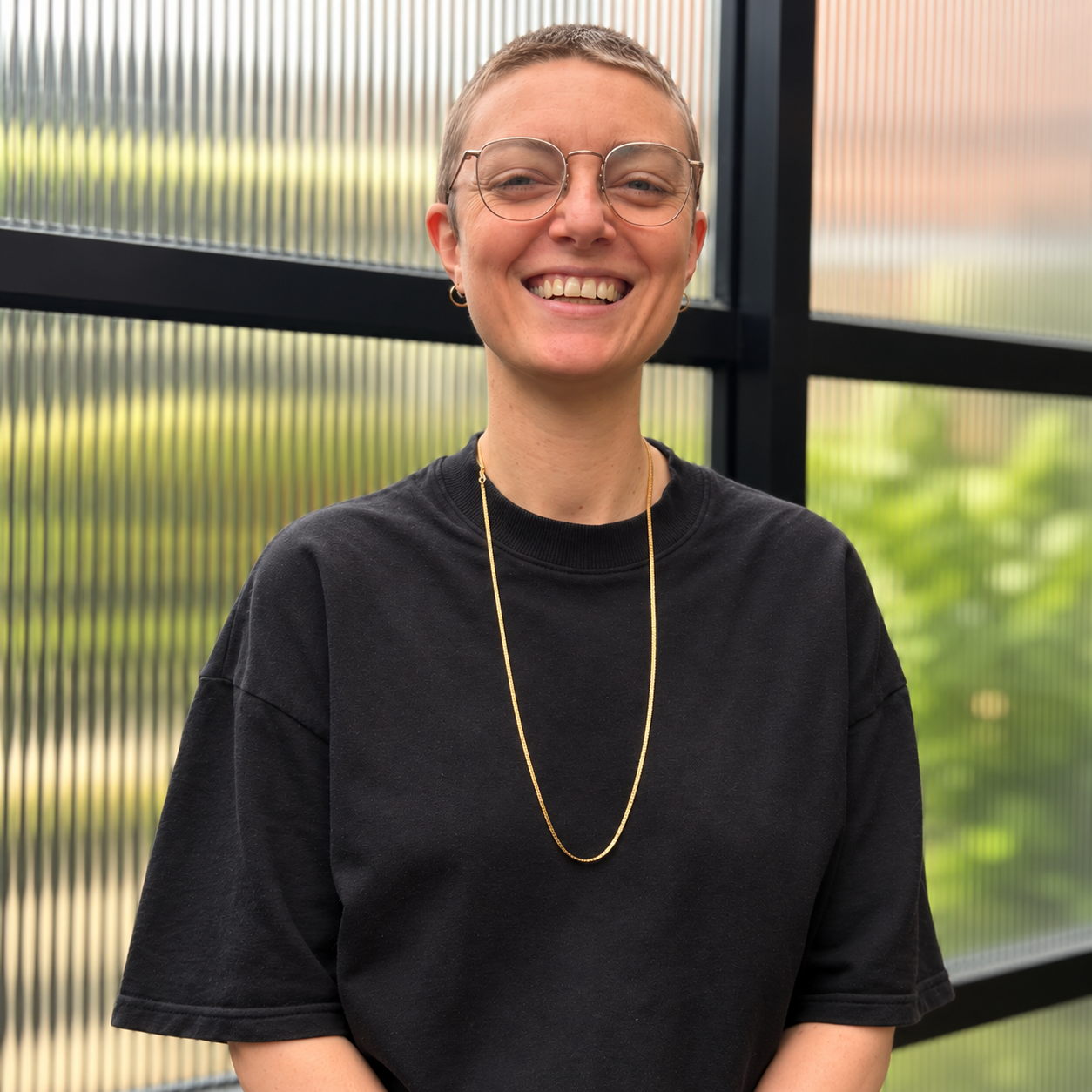 Close-up of a person with short hair, wearing glasses, earrings, and a black shirt, smiling in front of a corrugated metal background.