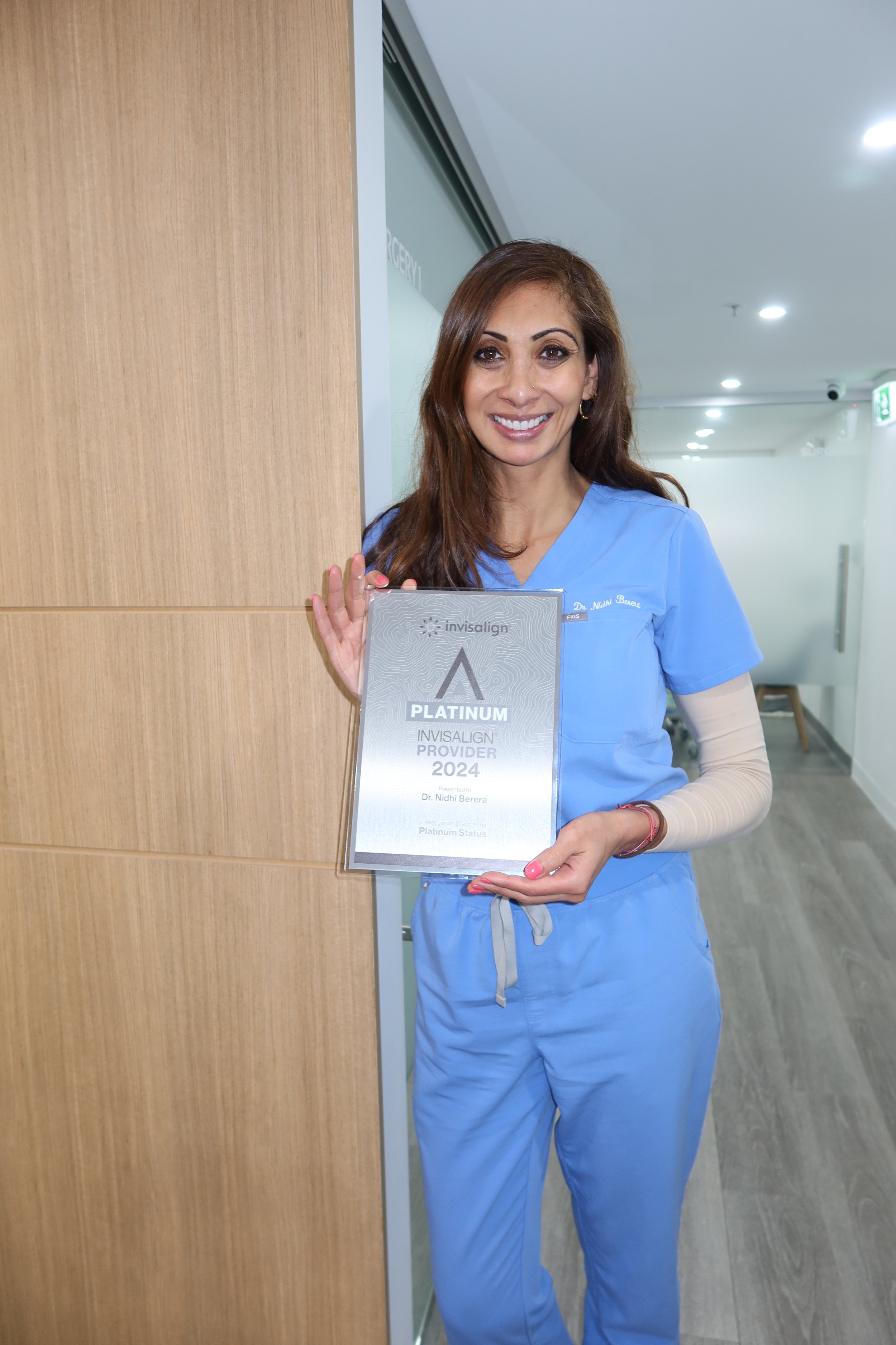 A woman in blue medical scrubs holding a plaque that reads 'Platinum Invisalign Provider 2024'.