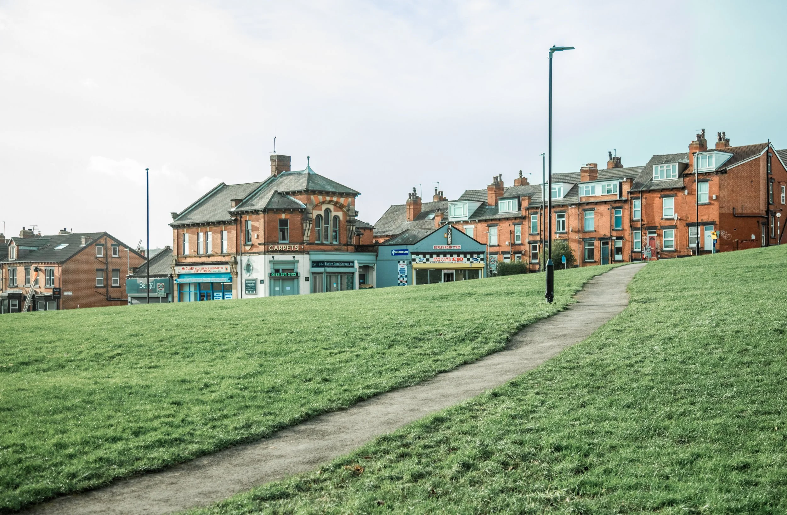 A grassy park with a dirt pathway, lamp posts, and buildings in the background under a cloudy sky. burley hyde park leeds