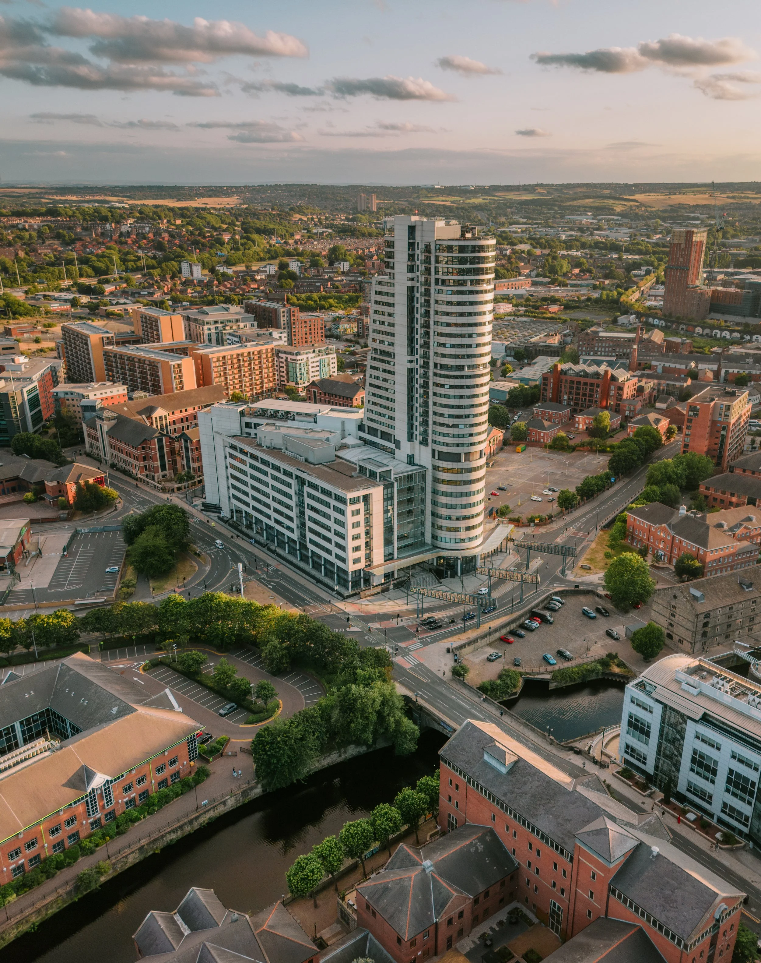Aerial view of a cityscape with modern high-rise buildings, historic brick buildings, a river, tree-lined streets, under a partly cloudy sky at sunset. bridge water place leeds cityc centre. drone photography leeds.
