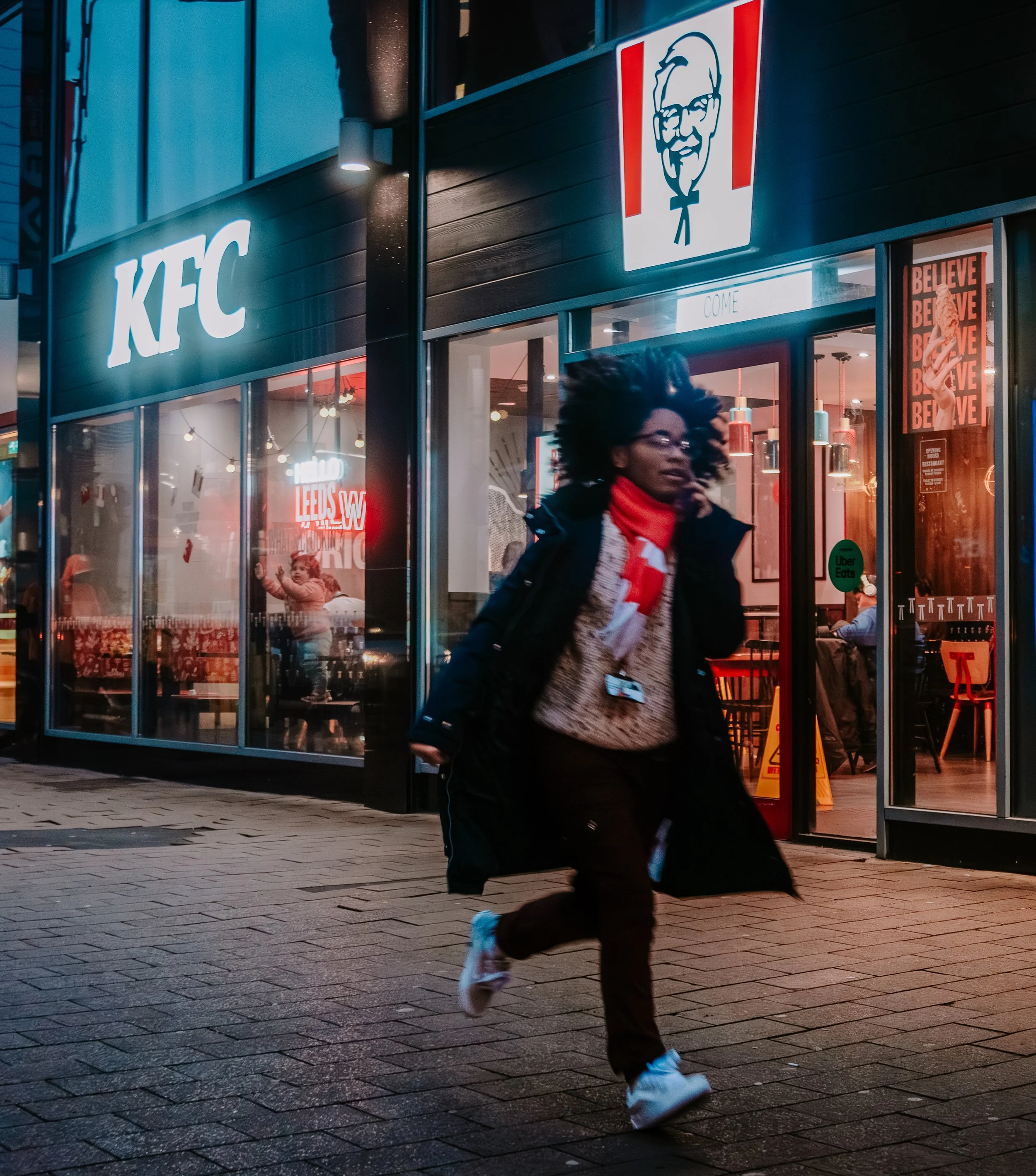 A woman walking a black and white dog outside a KFC restaurant at night. The woman has curly dark hair, glasses, a red scarf, and a black winter coat. The restaurant has bright interior lighting, with red and white decor, and visible signage includin
