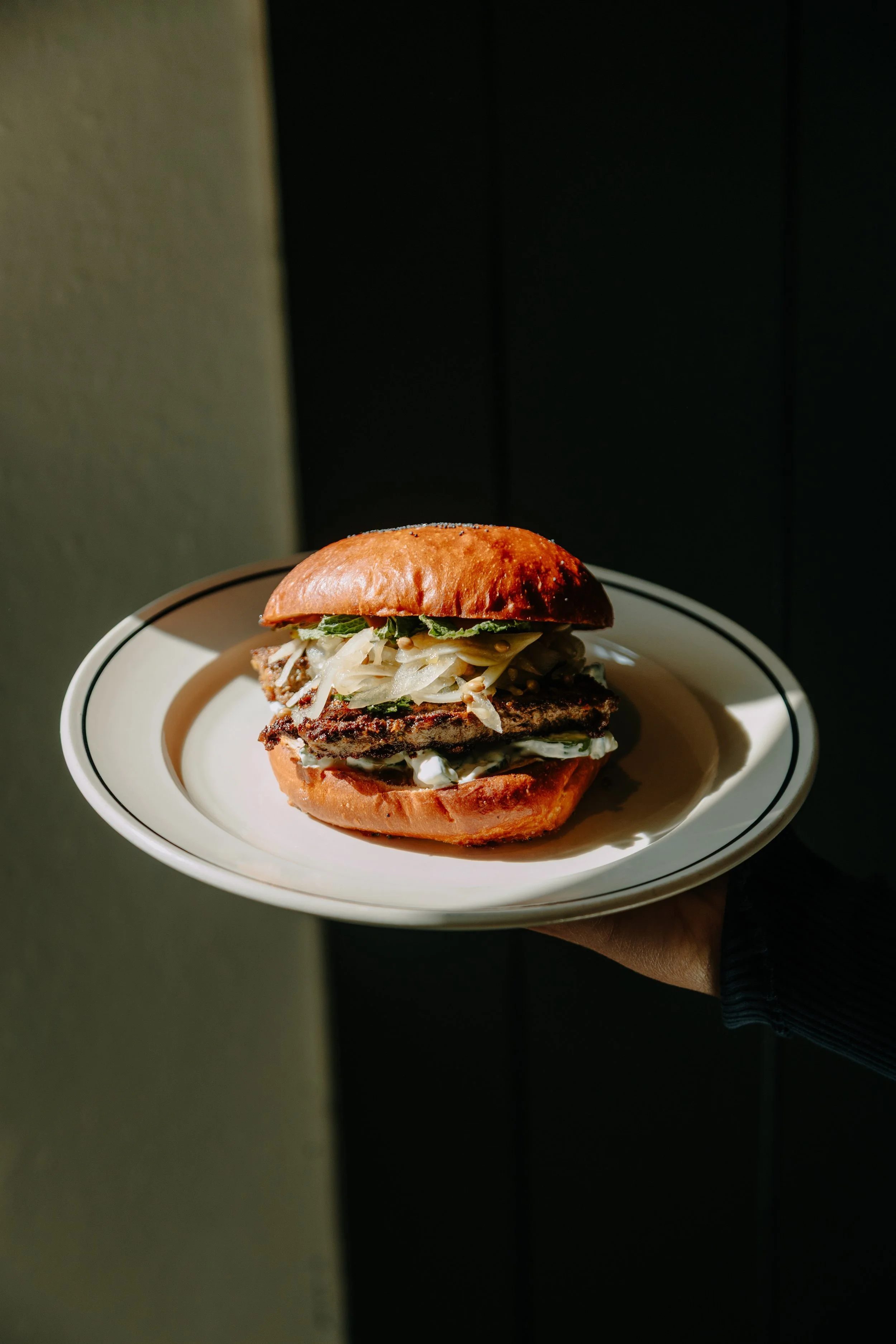 A burger with a toasted bun, topped with lettuce, shredded cheese, and a grilled beef patty, held on a white plate with black lines around the rim, against a dark background. york leeds yorkshire restaurant photographer videographer