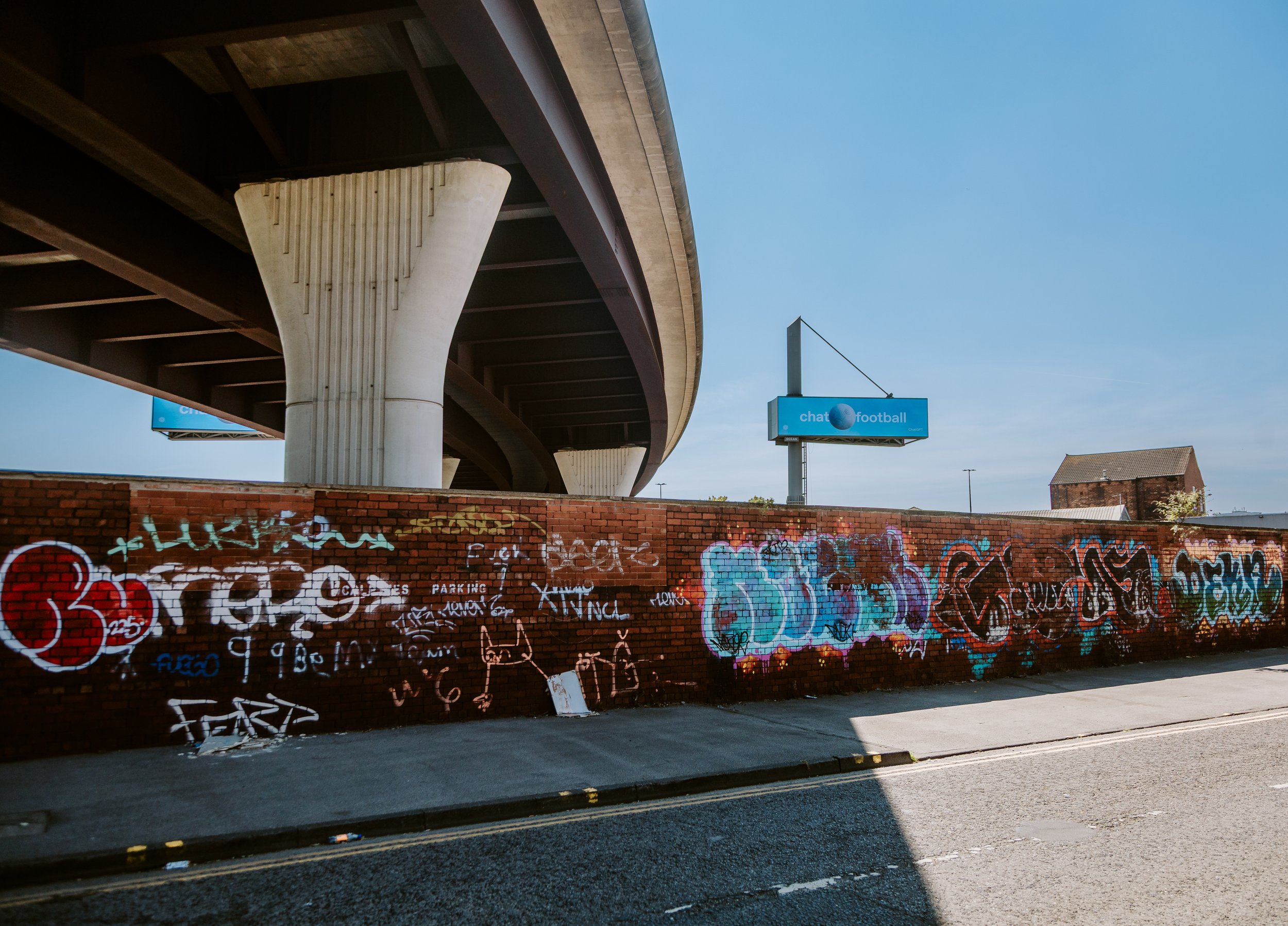 A street scene with a graffiti-covered brick wall in the foreground and a large elevated highway above. There are two billboards, one says 'chat football', and a roof of a building can be seen in the distance under a clear blue sky. Leeds hunslet