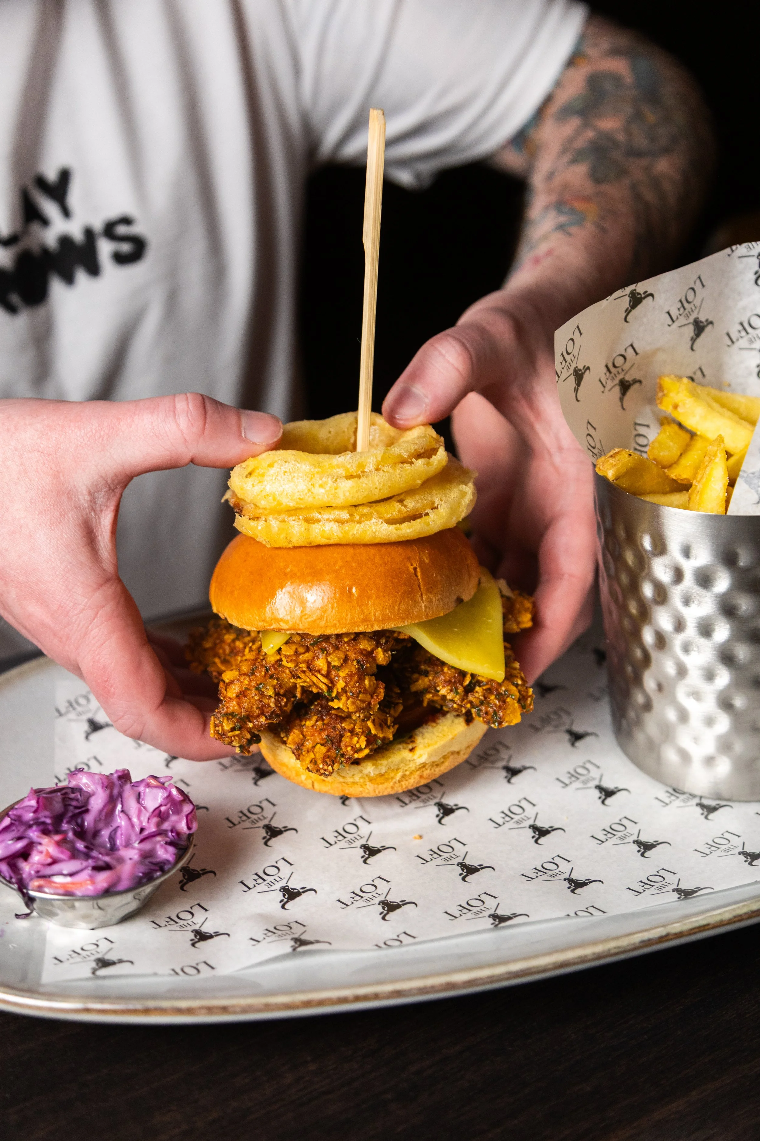 A person holding a fried chicken sandwich with a bun, pickles, cheese, and a spicy chicken fillet, with a side of French fries in a metal cup, and a small bowl of purple coleslaw on a tray lined with branded paper. The loft darlington. york leeds.
