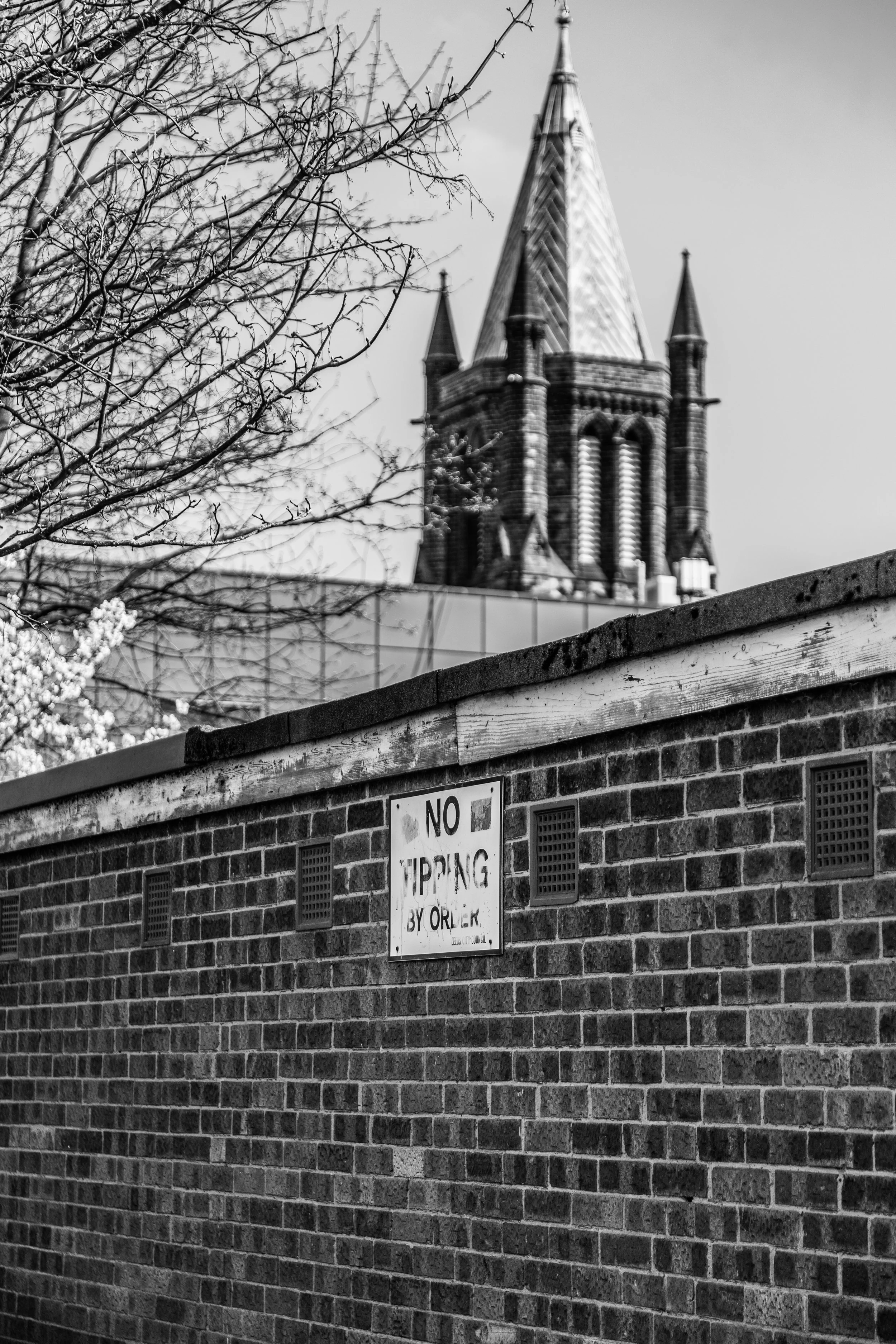 Black and white photo of a brick wall with a sign that says 'No Tipping by Order' in front of a church with a tall steeple and tree branches in the foreground.