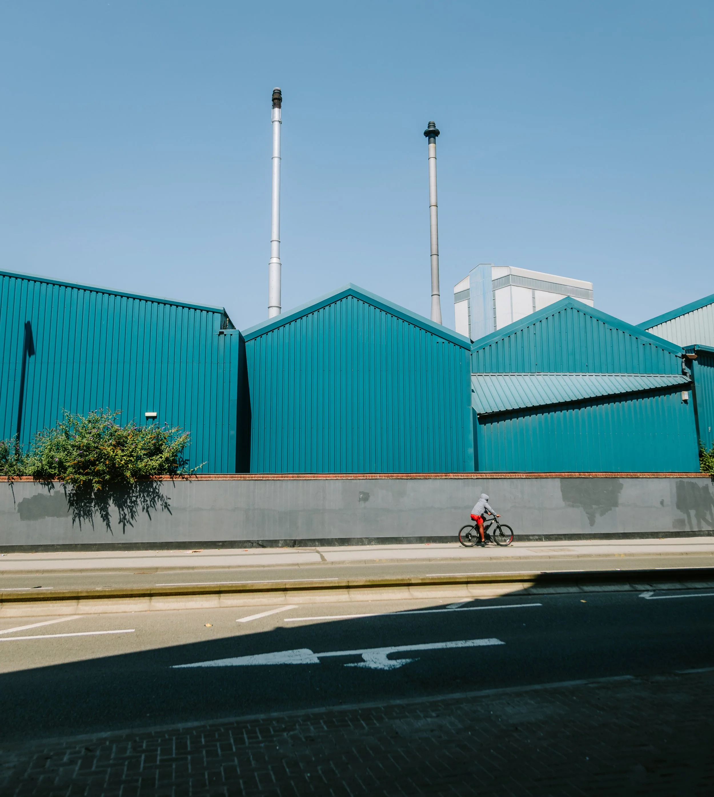 A person riding a bicycle along a city street in front of a large building with teal-colored corrugated metal walls and two tall industrial chimneys against a clear blue sky. Hunslet leeds.