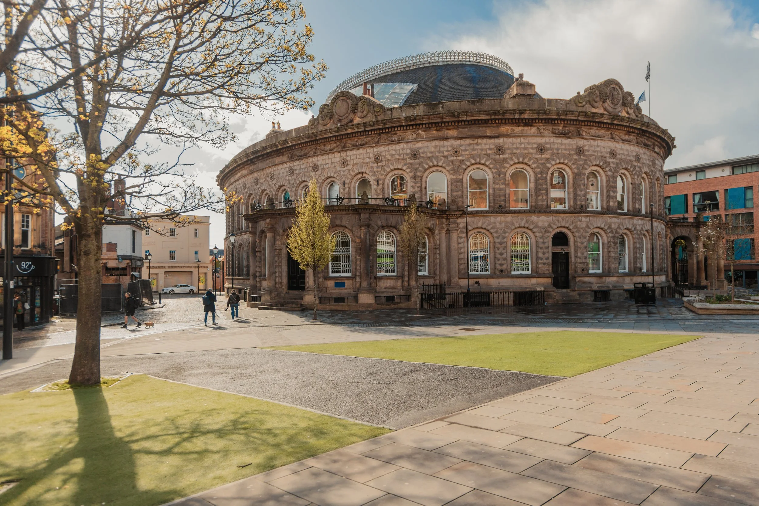 A historic round stone building with decorative architectural details and arched windows, situated in a park with a cobblestone street, trees, and a few people walking, under a partly cloudy sky. corn exchange leeds.