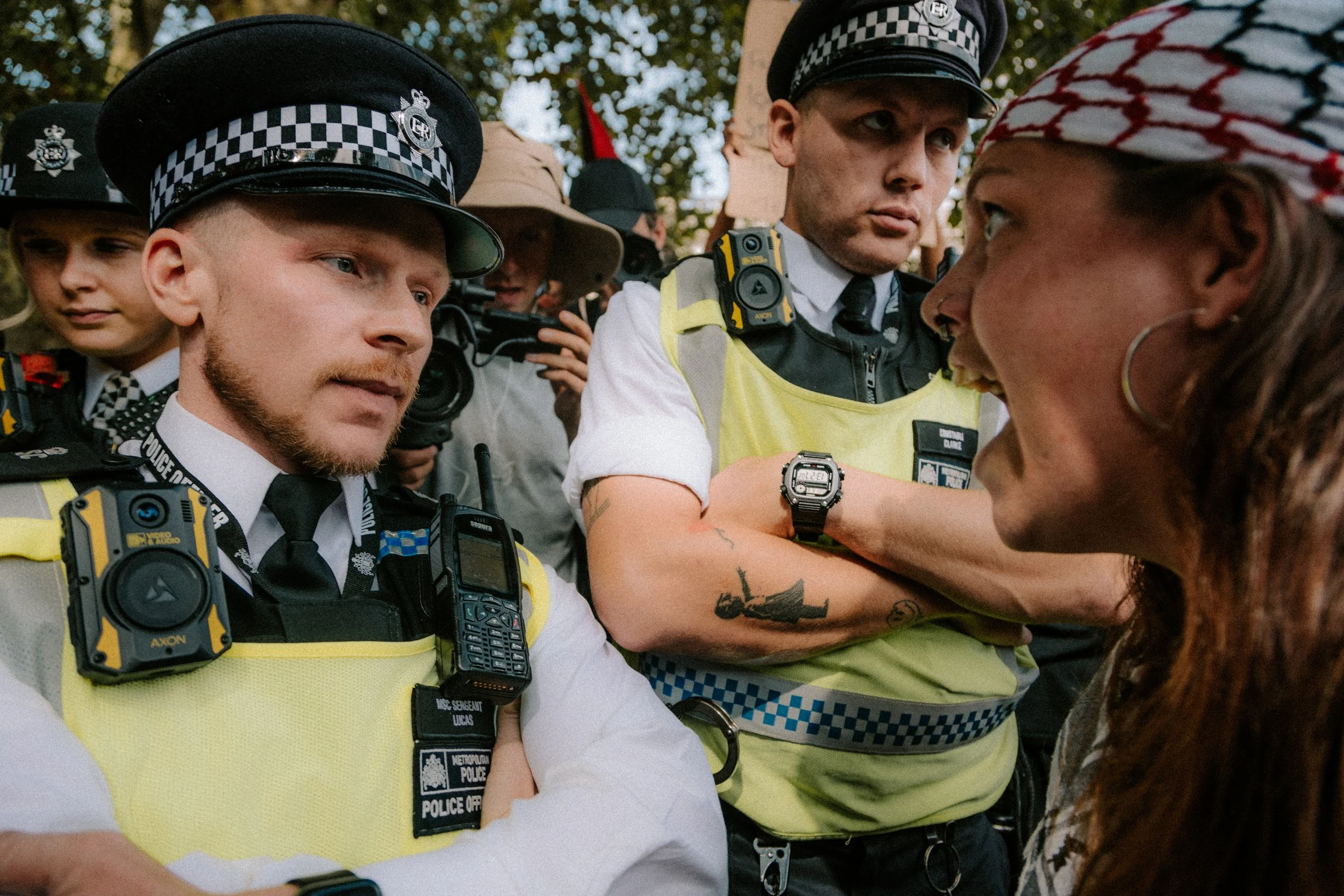 Police officers in uniform and reflective vests arrest a woman with long, curly hair and a patterned headscarf during a protest or demonstration, with other protesters in the background.