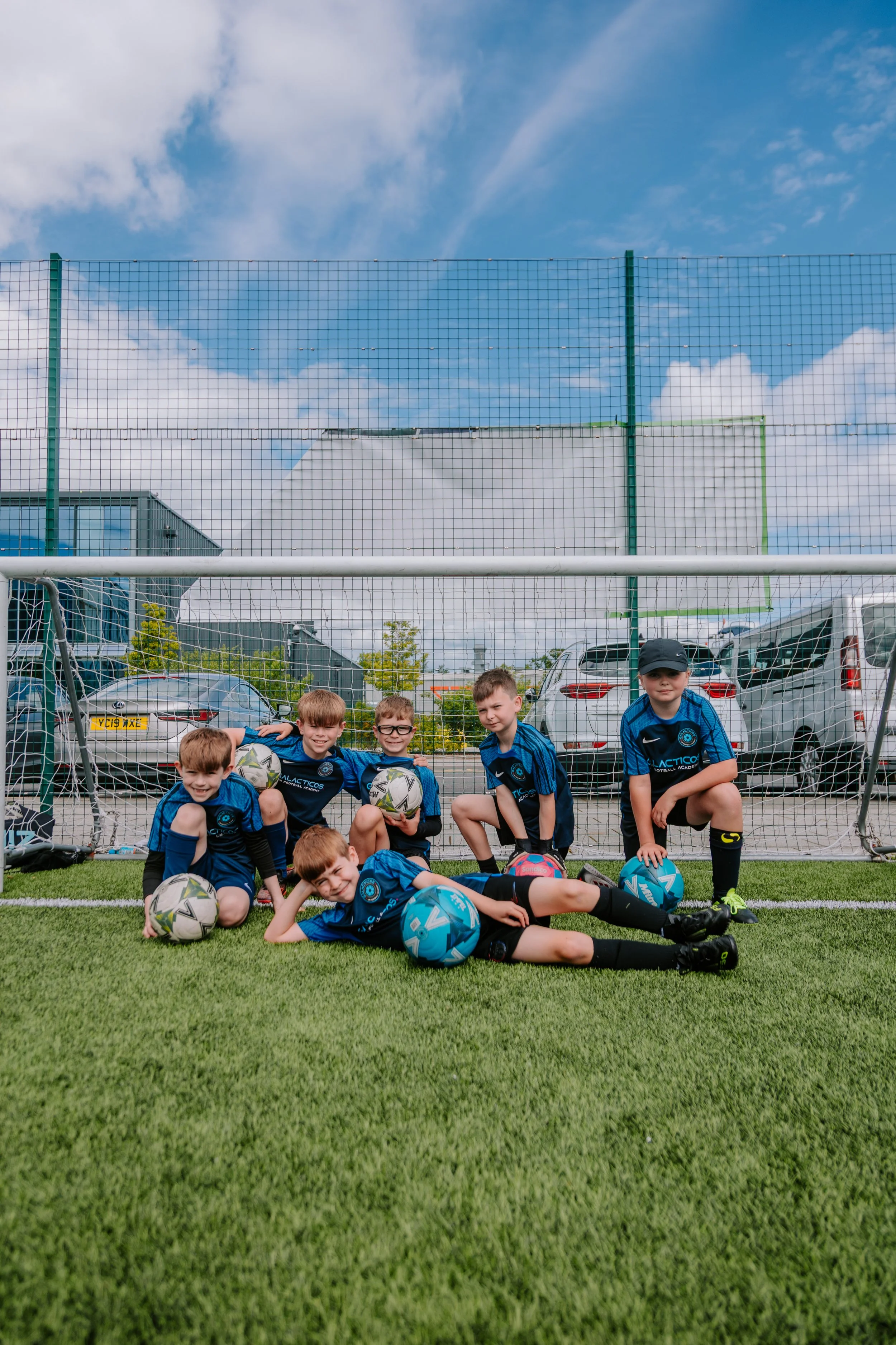 Group of young boys in blue soccer uniforms on a soccer field, posing with soccer balls after a game or practice, in front of a soccer goal and parked cars on a sunny day. sports photography
