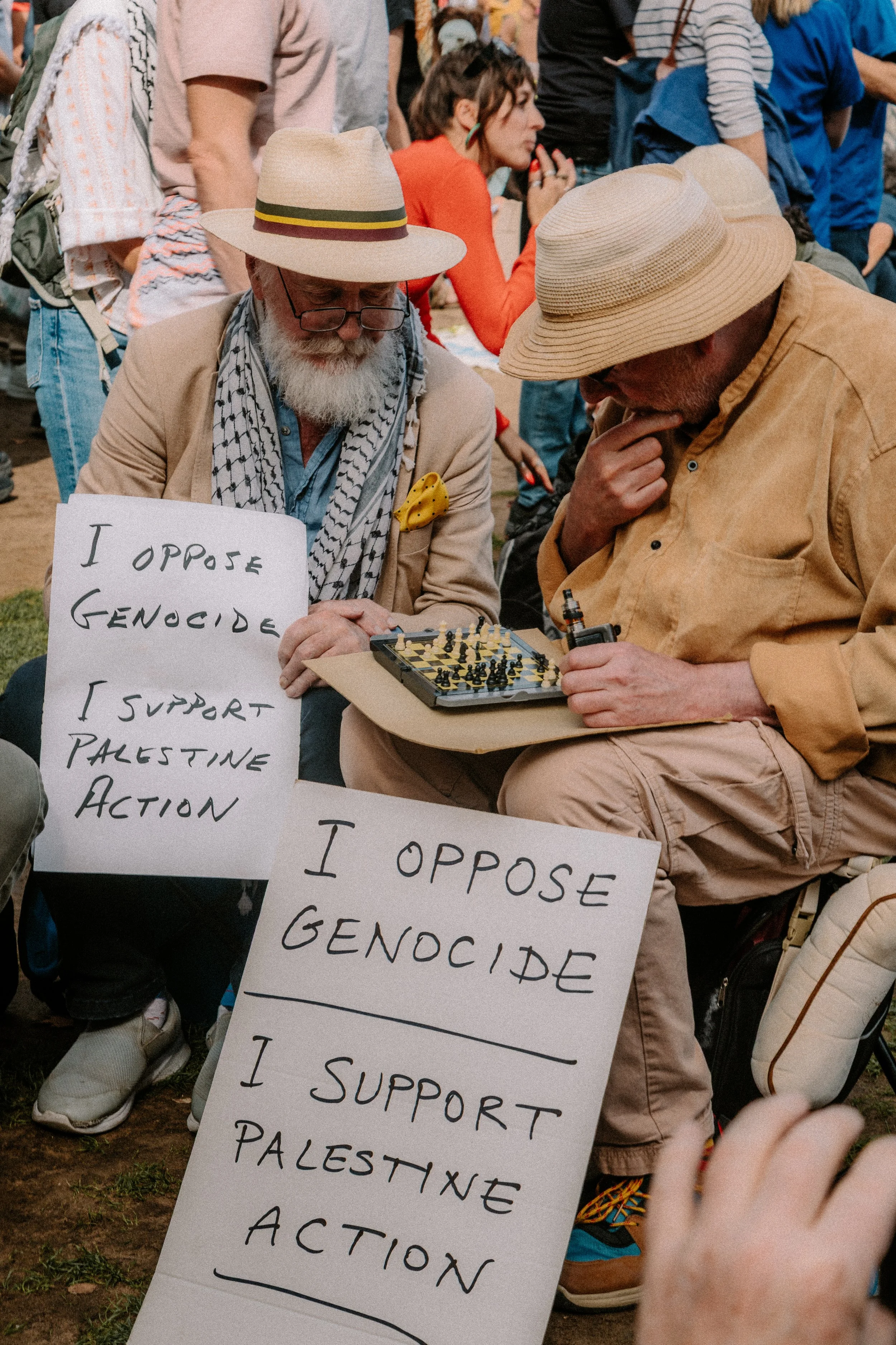 People gathered at a protest, holding signs that oppose genocide and support Palestine Action, with some individuals playing chess.