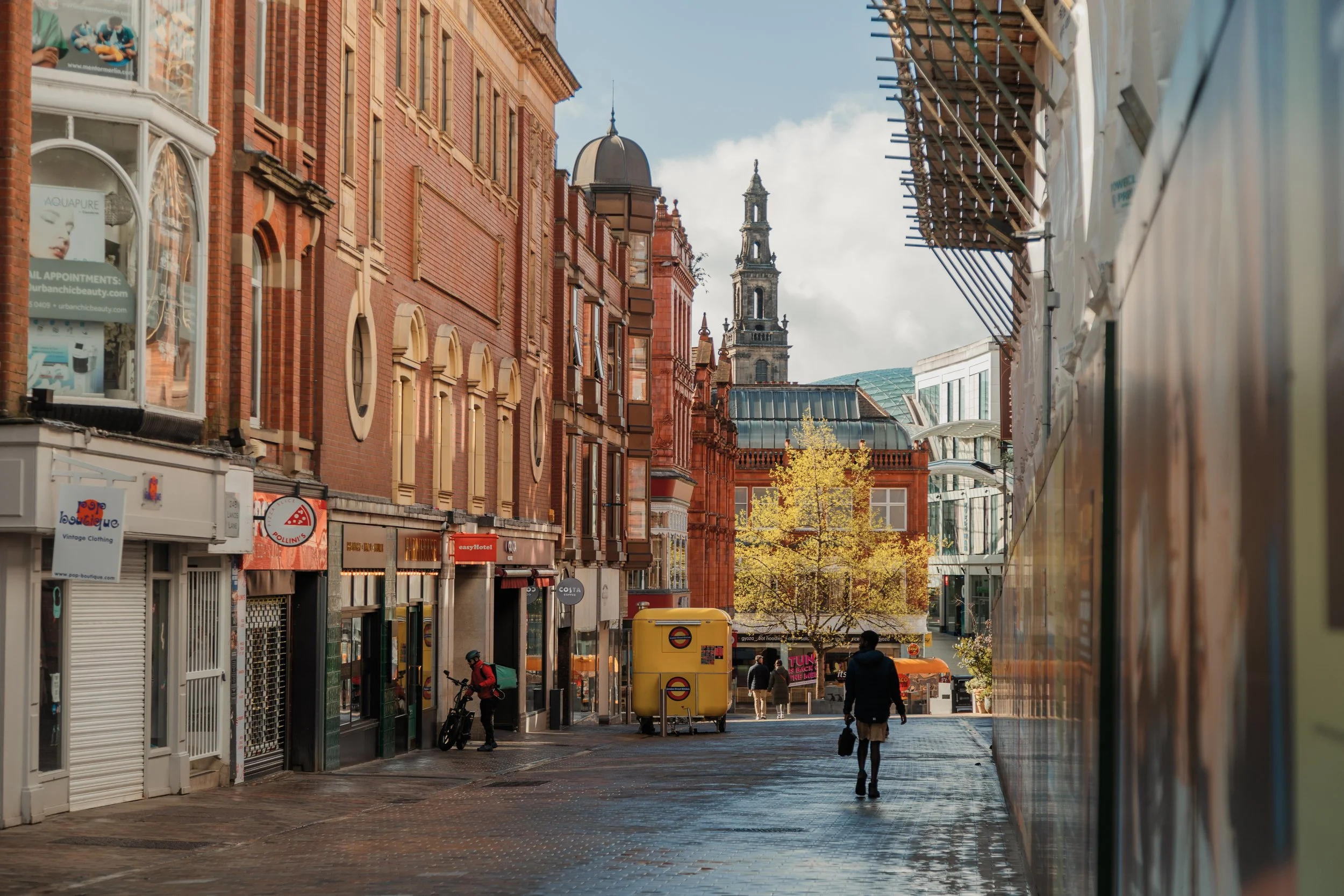 City street scene with red brick buildings, storefronts, pedestrians, and a tree with yellow leaves, with a church tower in the background. Leeds city centre, trinity shopping centre.