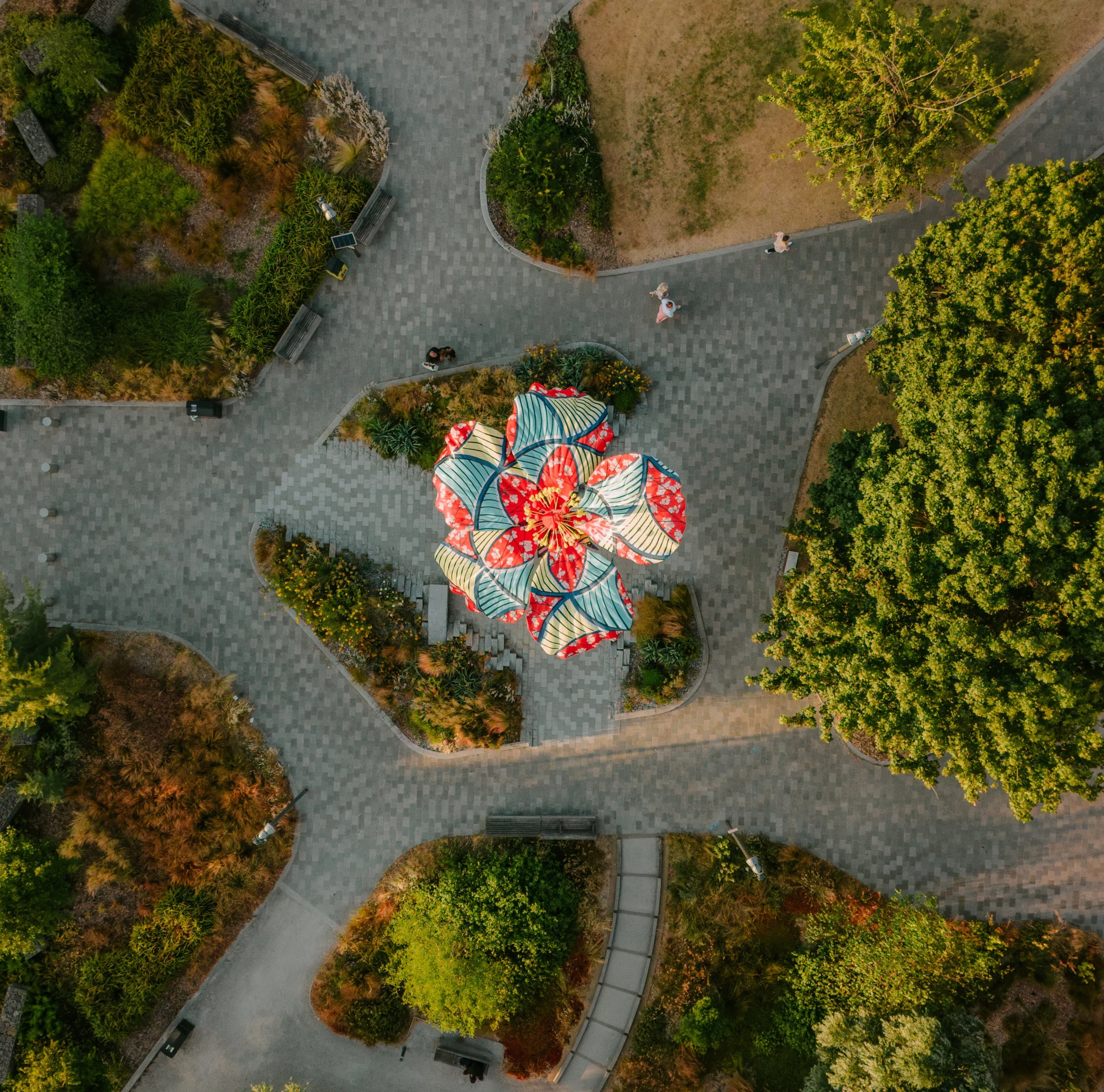 An aerial view of a park with paved walkways, green trees, benches, and a person walking. There are colorful umbrellas at the bottom of the image. hibiscus rising leeds city centre aire park.
