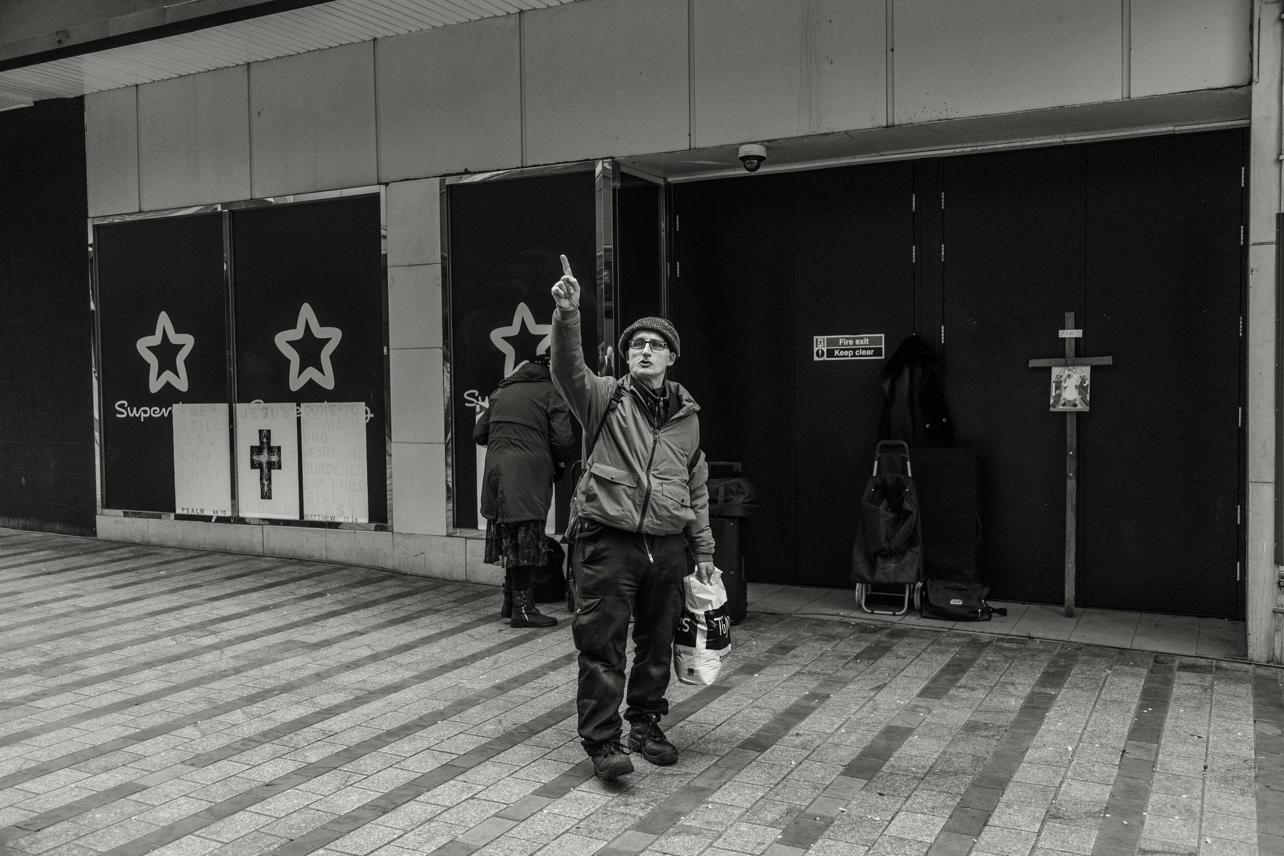 A man wearing glasses, a beanie, and a jacket is standing on a sidewalk and pointing upward with his right hand. In the background, there are shops with signs that include a star logo and a cross sign, along with a woman standing behind the man and s