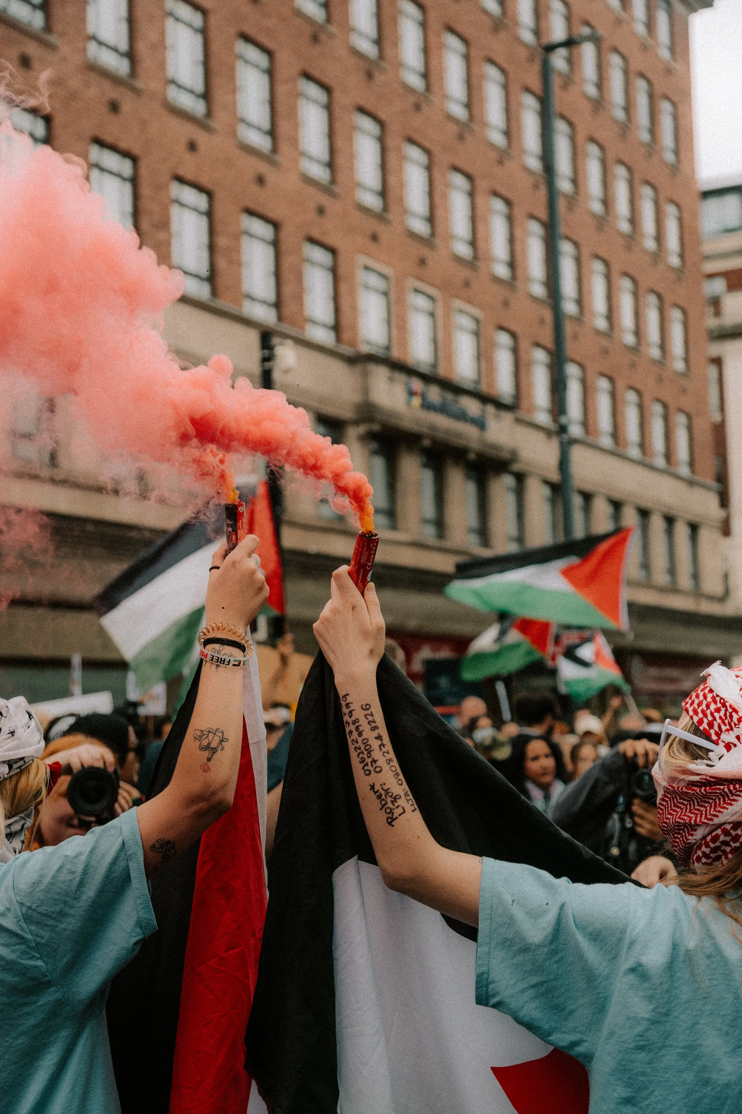 People holding pink smoke bombs during a public gathering in an urban street. The crowd is waving flags with a red, white, green, and black design, and there are flags draped over some of the people. A large brick building is visible in the backgroun
