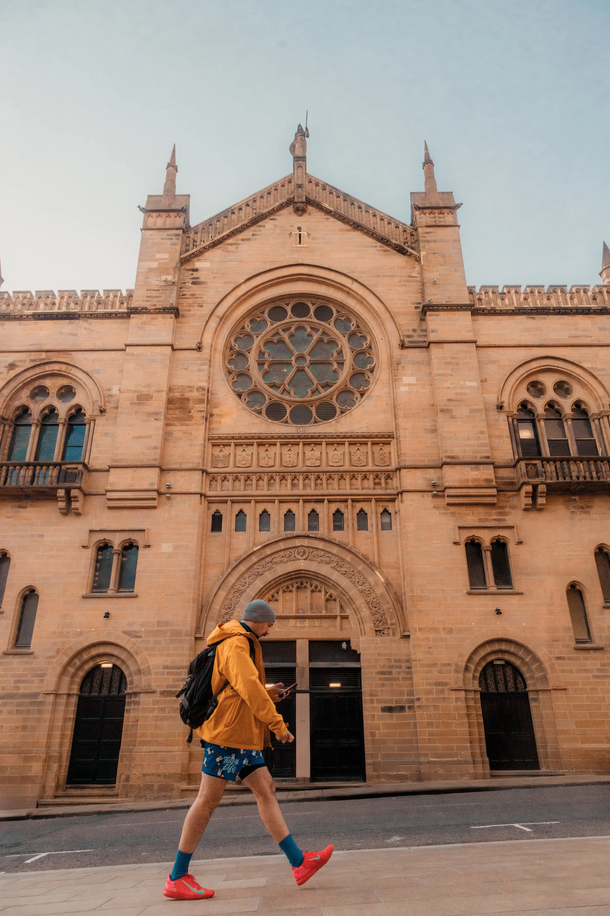 A man dressed in a yellow jacket, blue shorts with cartoon characters, bright red sneakers, and blue socks, walking past a large historic church with a rose window and intricate stone carvings in the early morning or evening. Leeds City Centre
