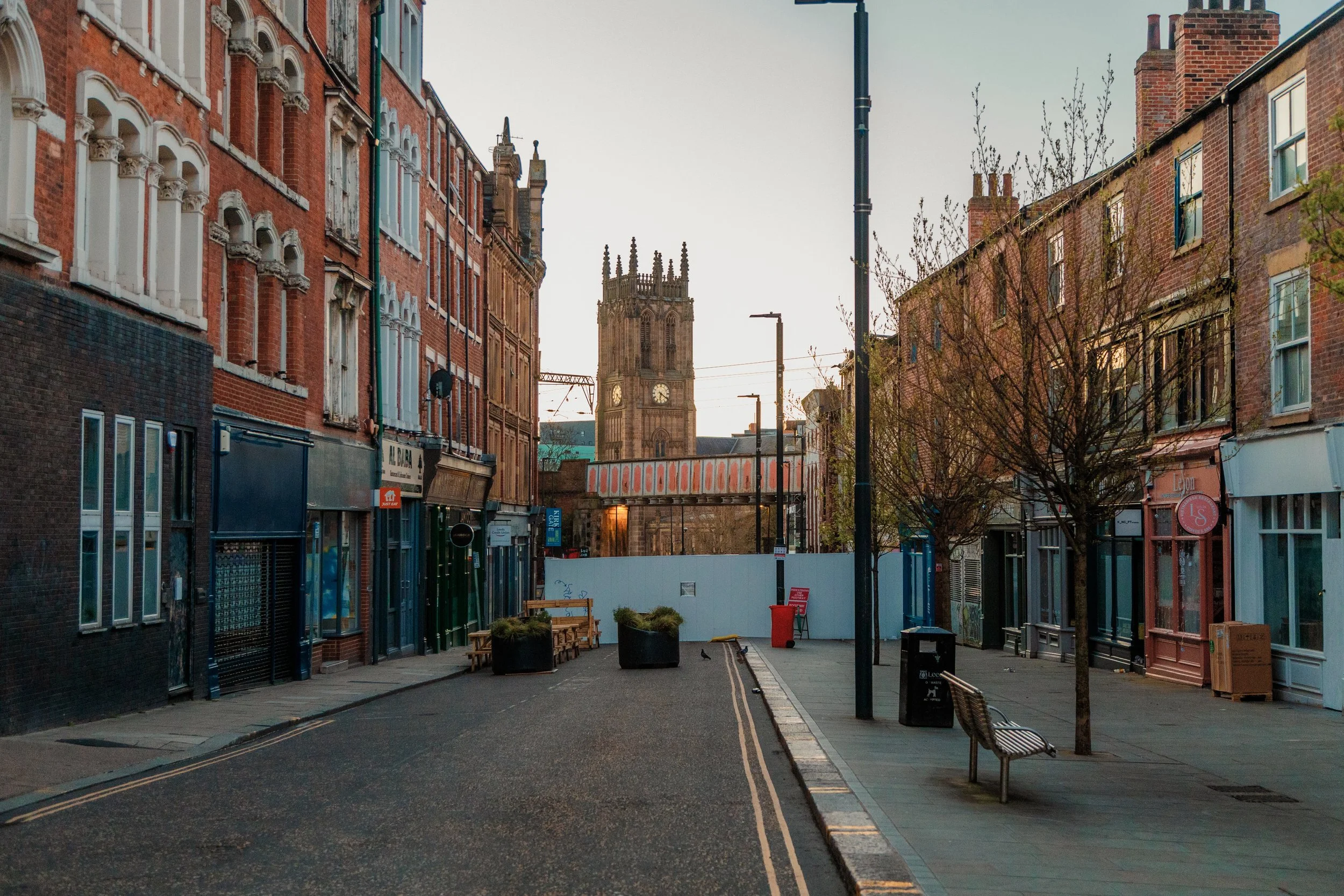 Empty city street with red brick buildings on either side, a church tower in the background, leafless trees, benches, streetlights, and planters, during daytime. Leeds minster.