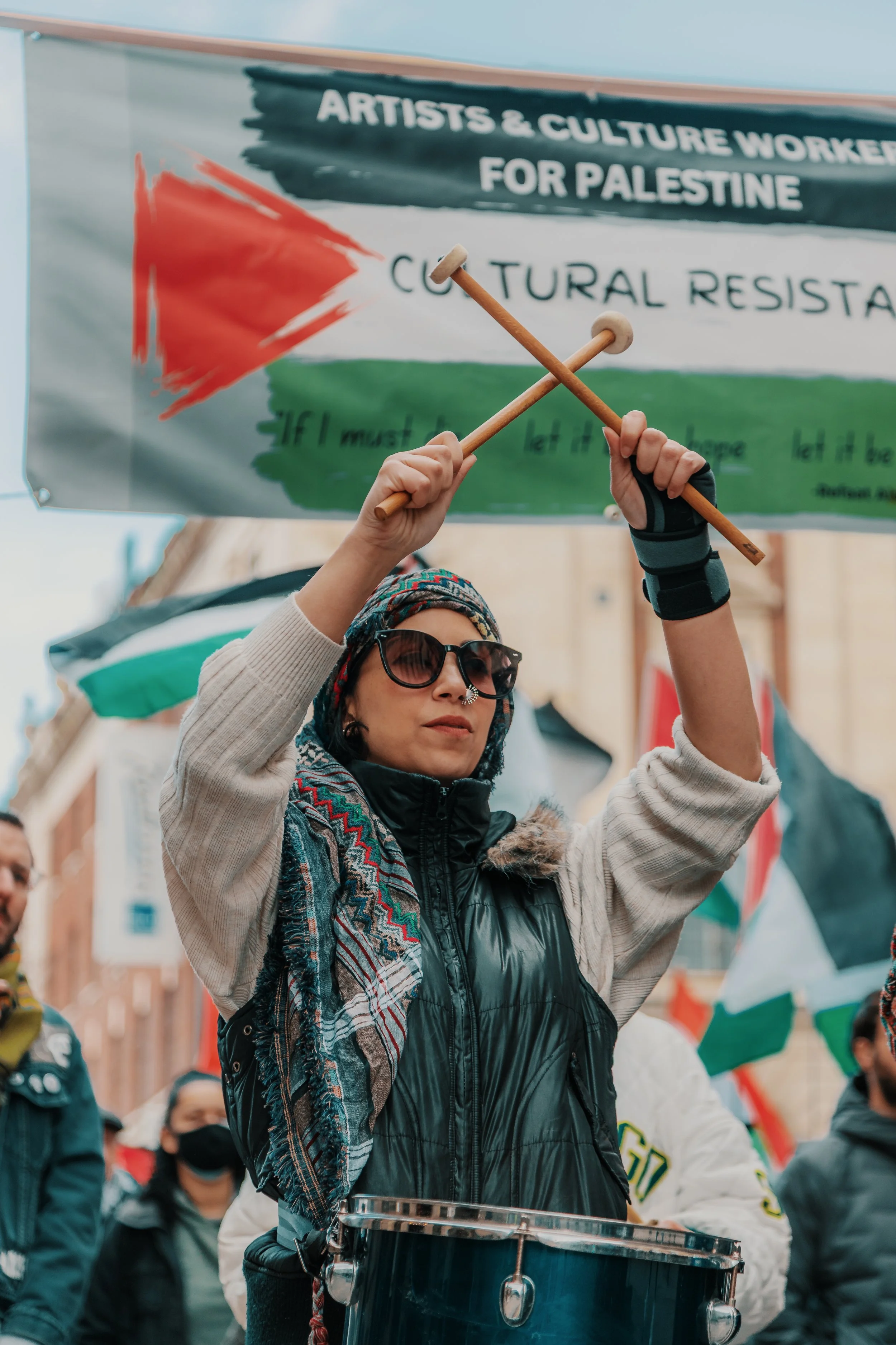 A woman wearing sunglasses, a headscarf, and a vest, holding drumsticks and playing a drum in a crowd during a protest. A banner in the background reads 'Artists & Culture Workers for Palestine'.