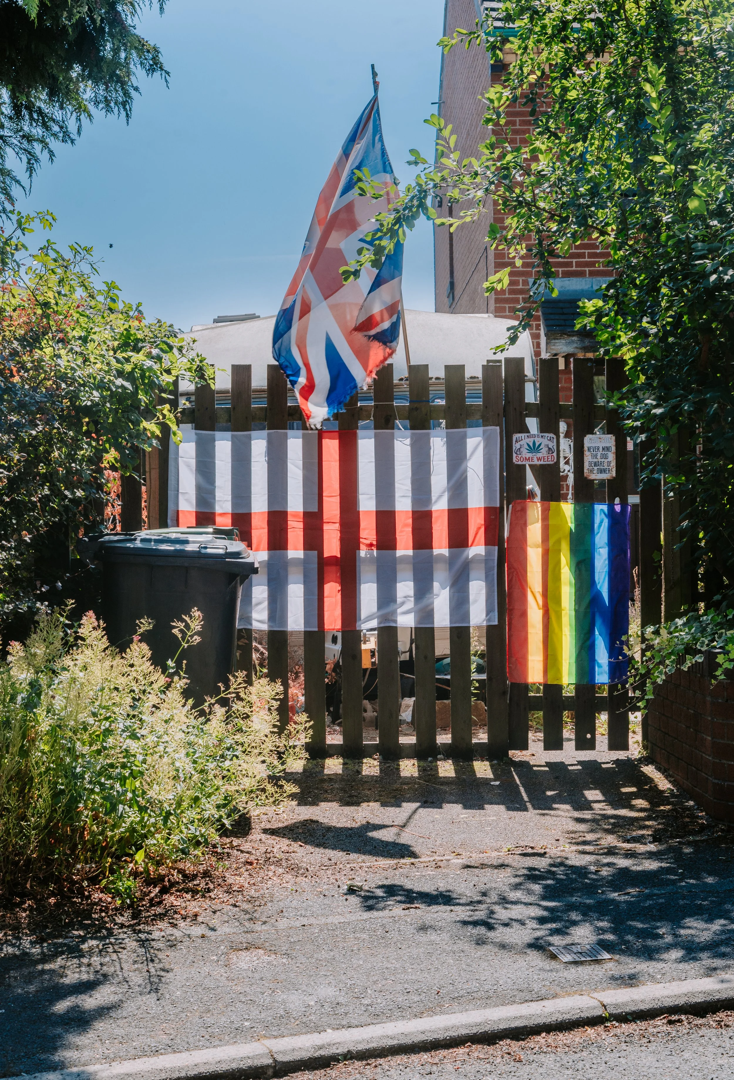 A neighborhood backyard gate decorated with rainbow flags, a Union Jack flag, and signs promoting marijuana and gay pride.