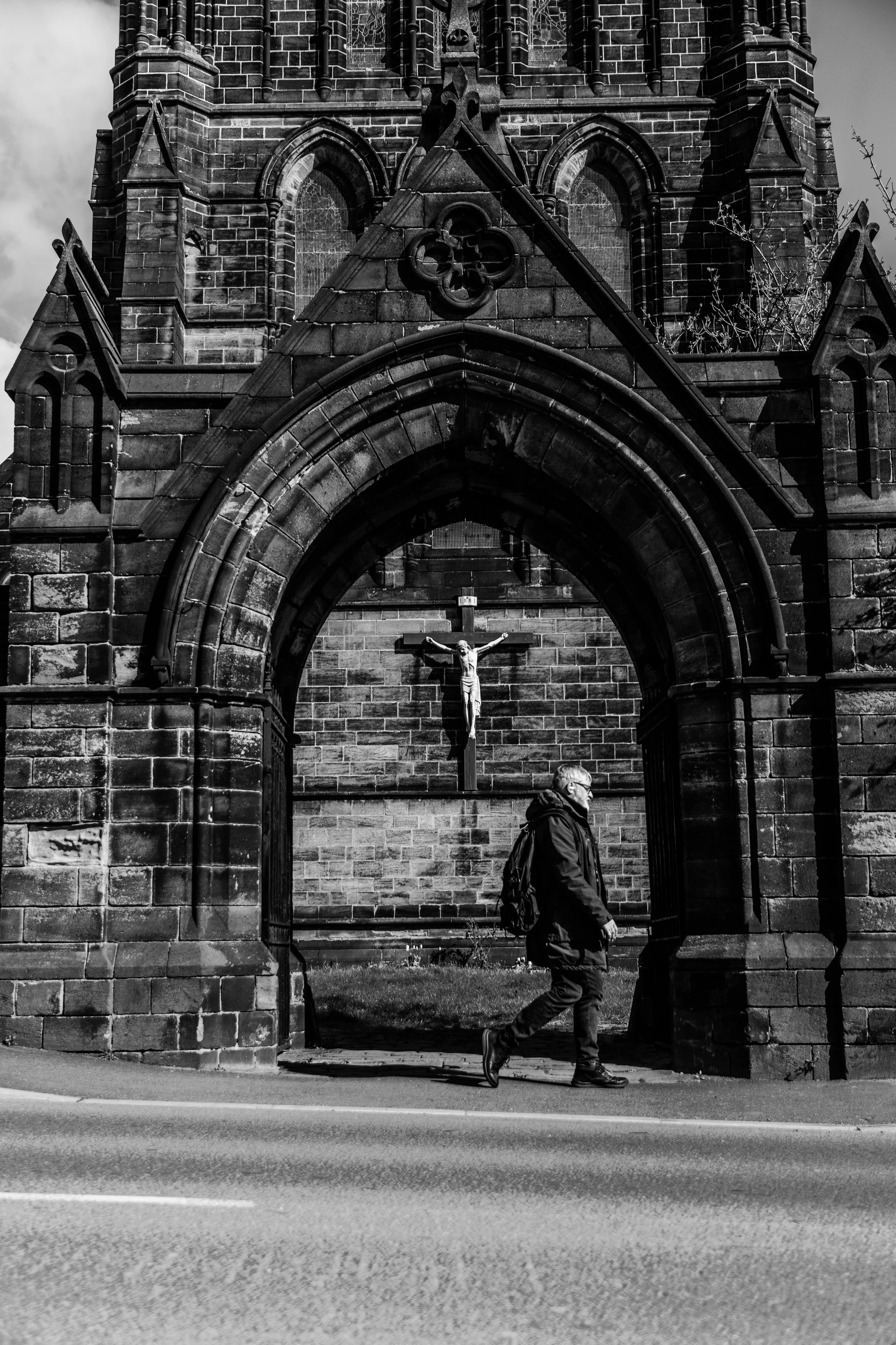 A man walking past a stone church with gothic architecture, including arched doorways, windows, and a crucifix inside. The photo is in black and white, and a person with a backpack is in motion in the foreground.