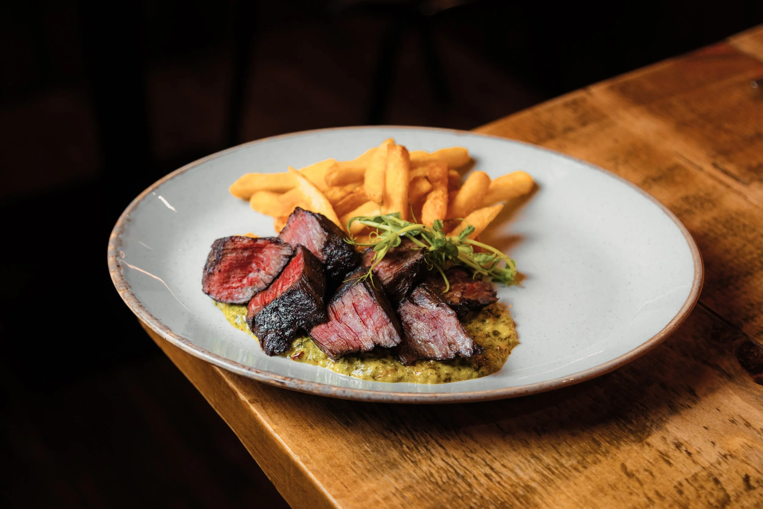 A plate of sliced medium-rare steak with a dark crust, served with crispy French fries and garnished with microgreens, on a ceramic dish resting on a wooden table. york leeds yorkshire restaurant photographer videographer