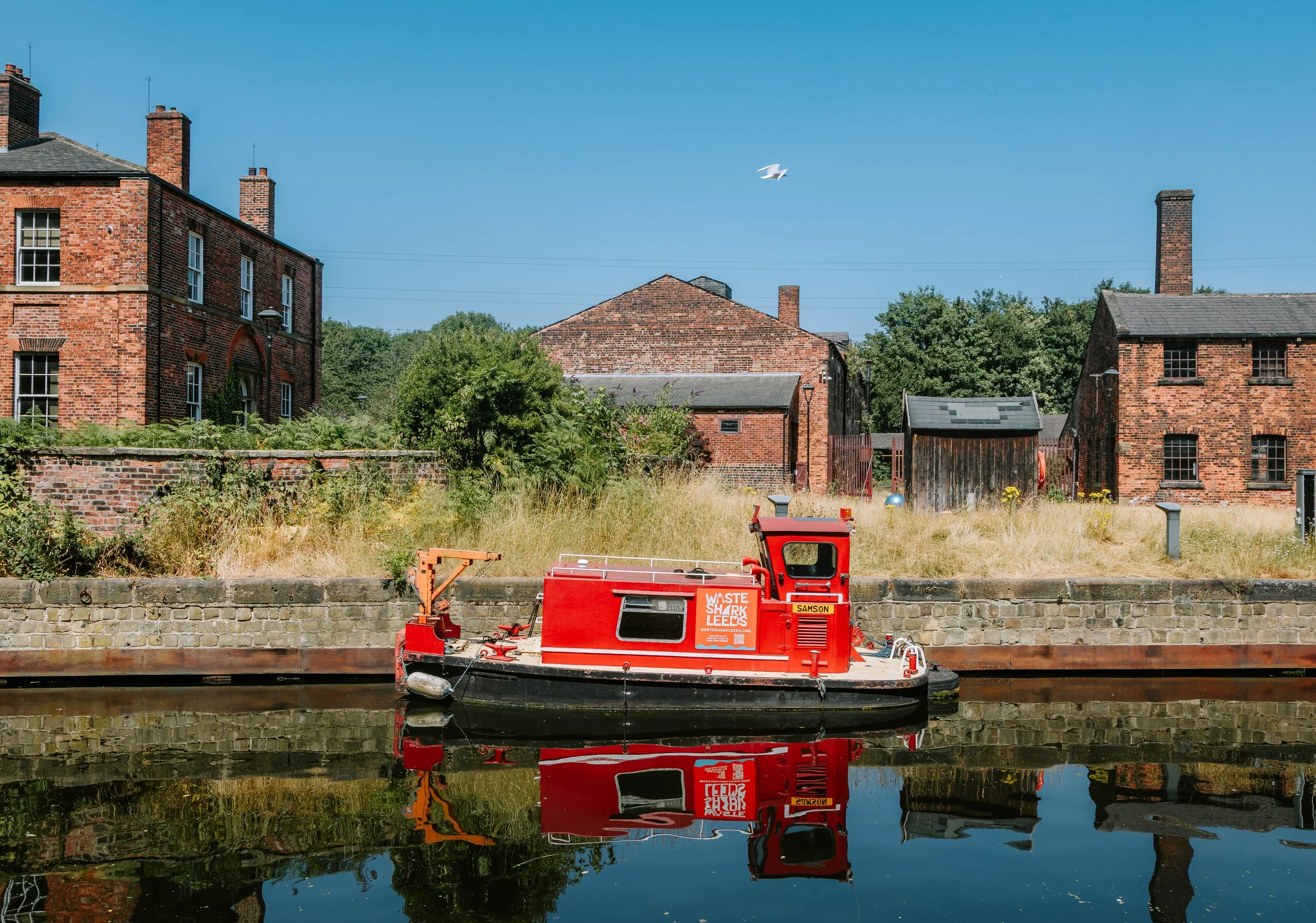 A red waste collection boat named 'Waste Shark Leeds' docked along a canal, with brick houses and greenery in the background under a clear blue sky. The boat's reflection is visible in the water.