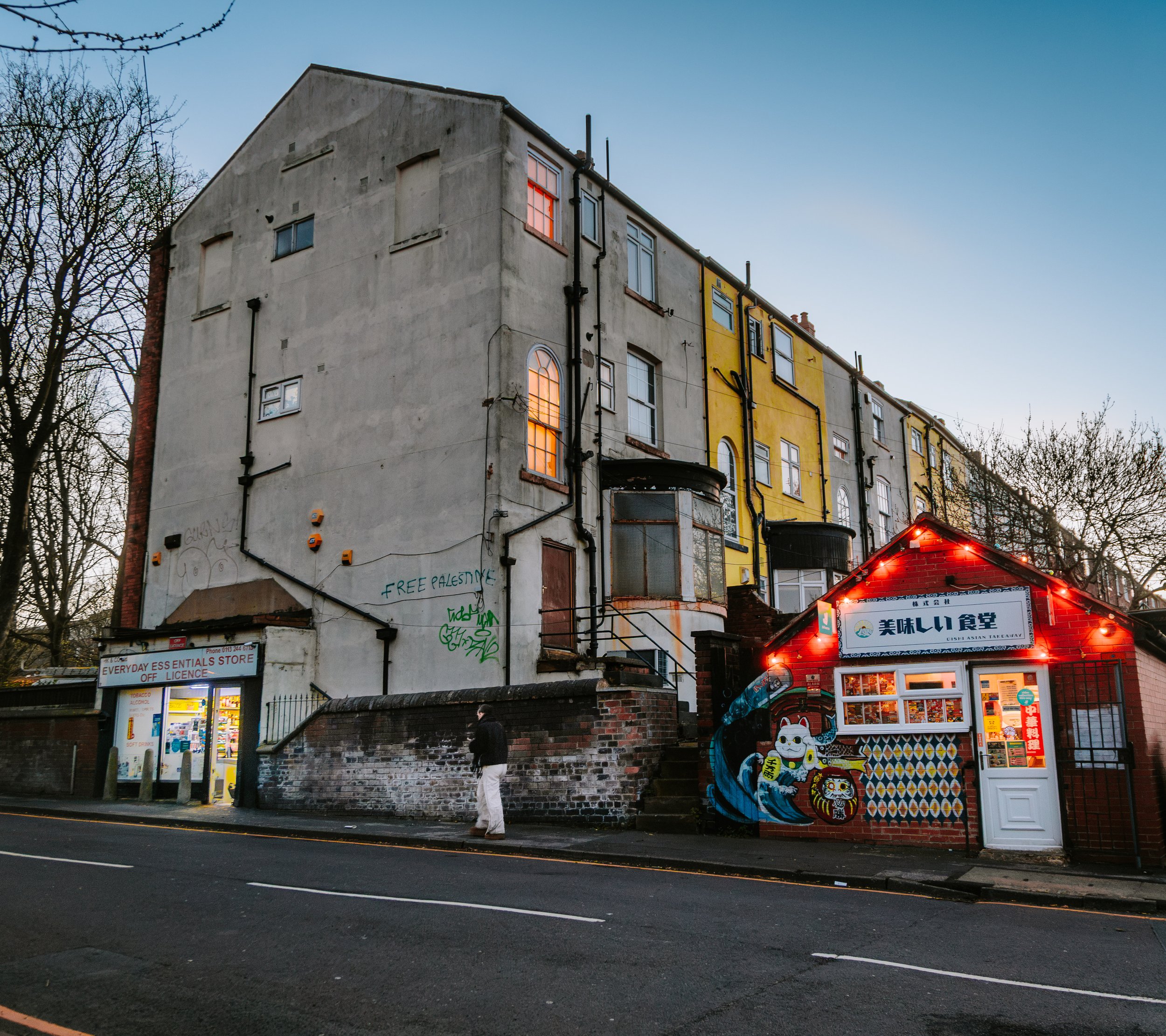 A multi-story building with a weathered exterior, some windows with lights on, and graffiti on the lower part, beside a street with a person walking and a small Asian food stand with bright red neon lights. Woodhouse leeds.
