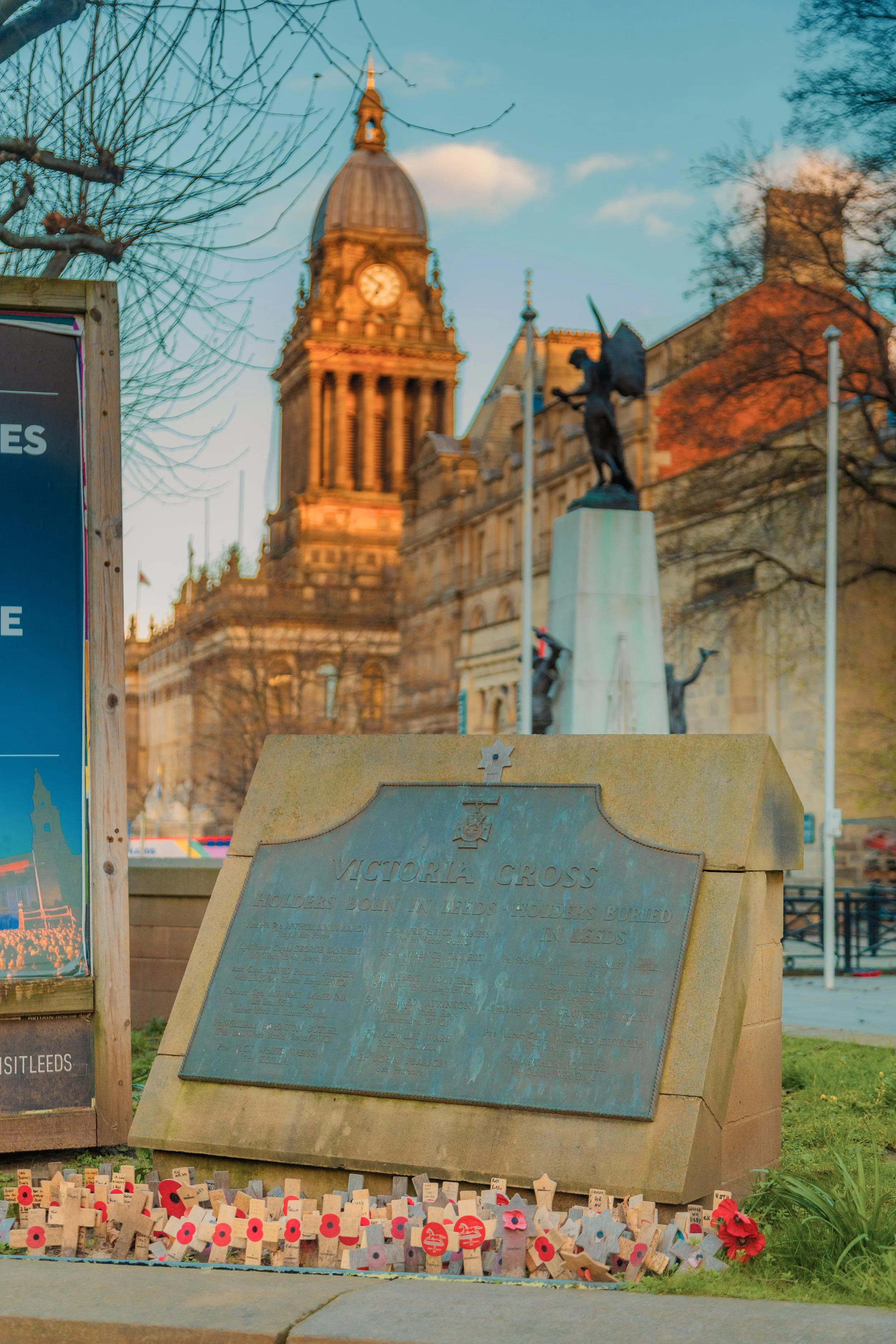 A memorial with a plaque dedicated to Victoria Cross recipients, surrounded by small poppy crosses. In the background, a clock tower and a statue of a soldier holding a rifle. Leeds city centre, victoria square gardens, town hall