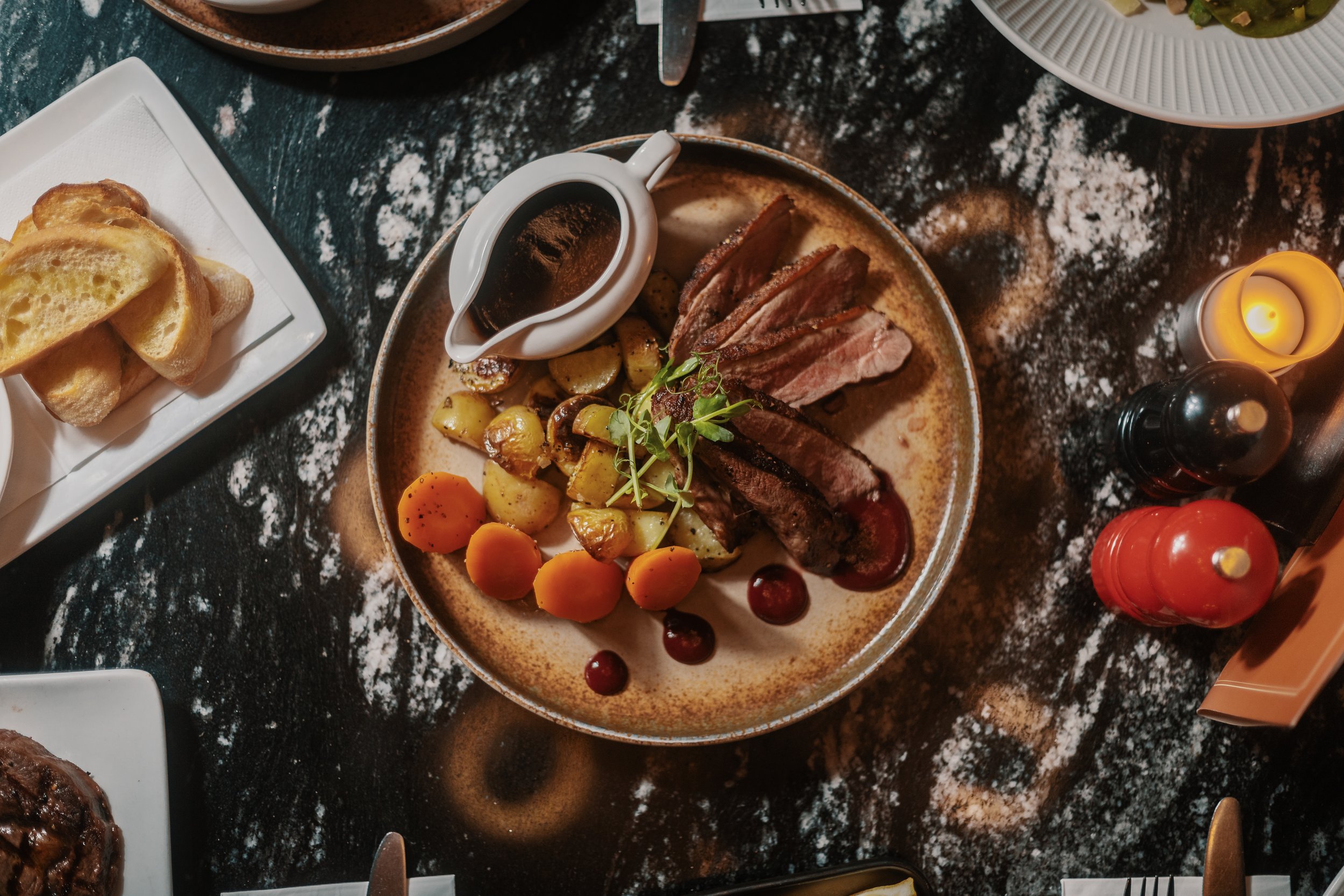 A plate with sliced beef, roasted potatoes, carrots, and green garnish served with gravy and sauce, on a table with other dishes, candles, and condiments. york leeds yorkshire restaurant photographer videographer