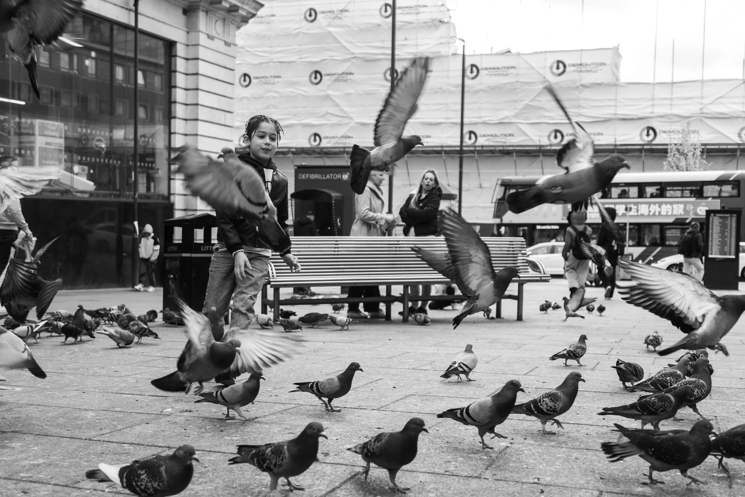 A young boy stands among pigeons in an urban city square, with pigeons flying and walking around. Behind him, women are standing near a wall with advertisements and a bus is visible in the background.