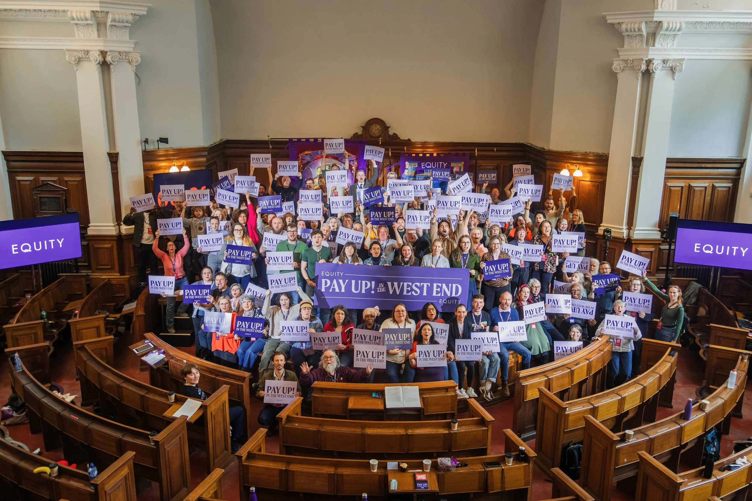 Group of people gathered in a large room, holding signs that read 'PAY UP! IN THE WEST END' and 'EQUITY'. The room has wooden paneling and ornate architectural details, resembling a historic or formal setting.