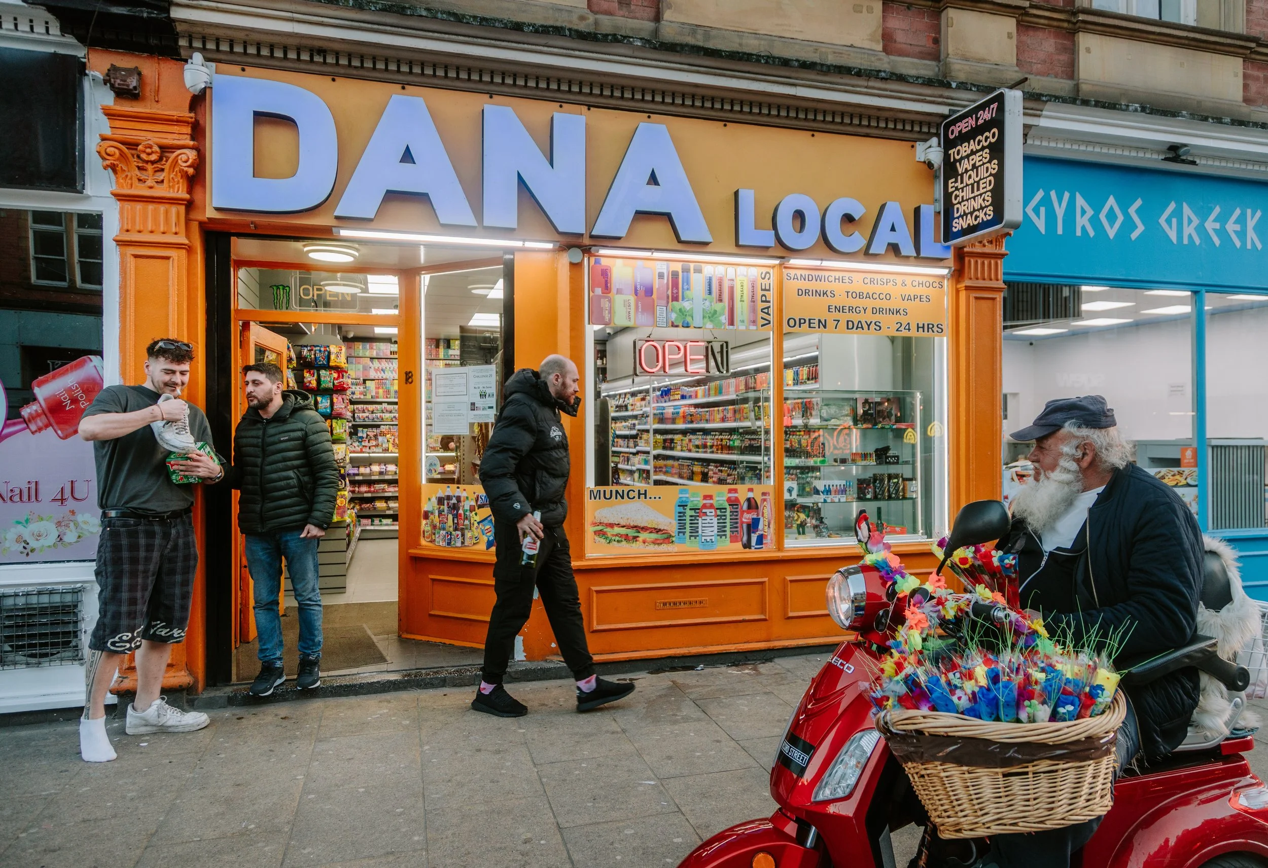 A small convenience store named 'DANA LOCAL' with bright orange exterior and large blue lettering, opening onto a street with pedestrians and a scooter decorated with colorful flowers in front. Leeds City Centre.