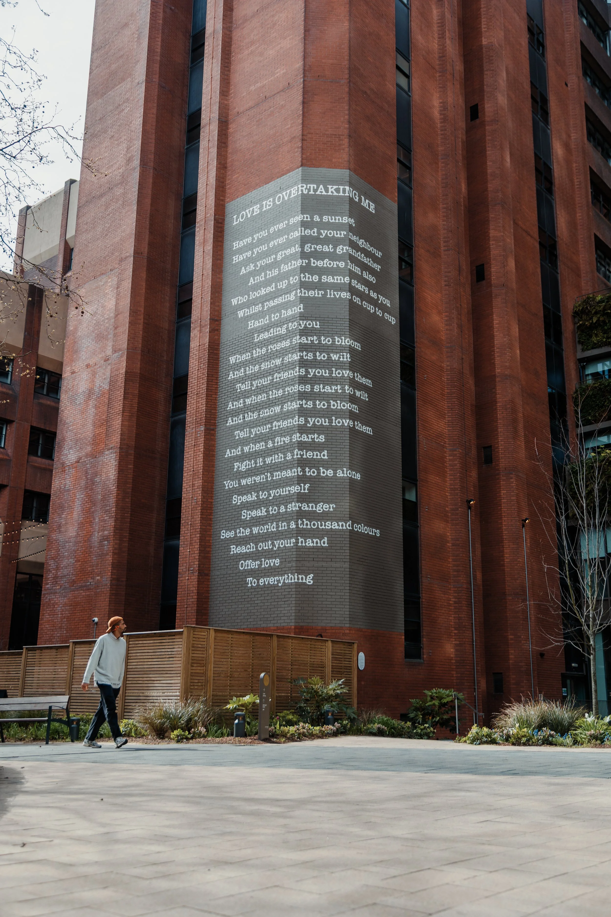 A large building with a high-rise brick exterior features a vertical mural of a poem painted on one side. In front, a man walks along a paved area near a garden with various plants and flowers, and a wooden fence separates the garden from the paved w