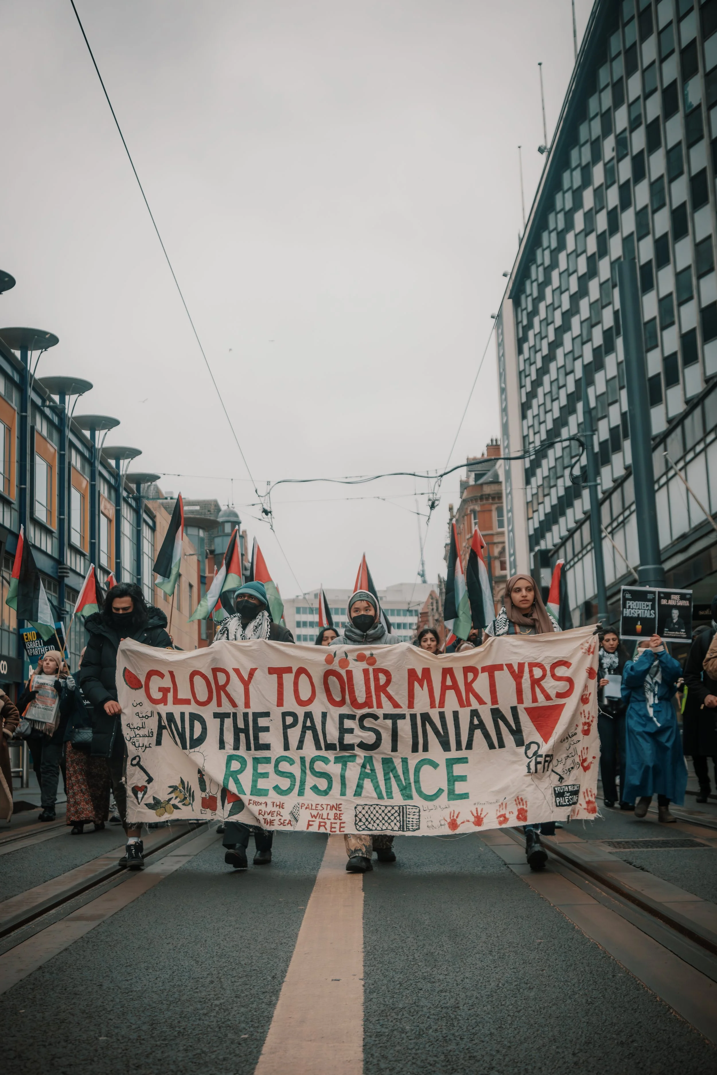 A group of protesters march down a city street, holding a large banner that reads 'Glory to Our Martyrs and the Palestinian Resistance.' The protesters are wearing face masks and winter clothing, with some waving flags. The buildings on either side o