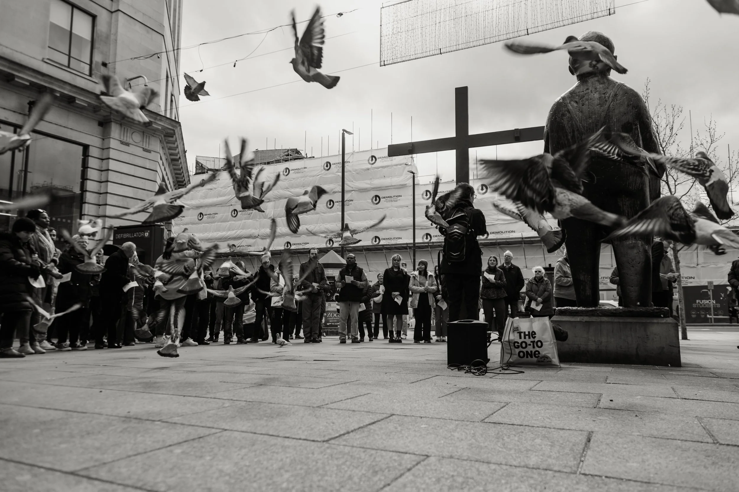 A black-and-white photo of a public gathering in an urban area with a statue of a religious figure, possibly the Virgin Mary, in the foreground. People are standing and watching a group of children or performers dressed in costumes, possibly for a da