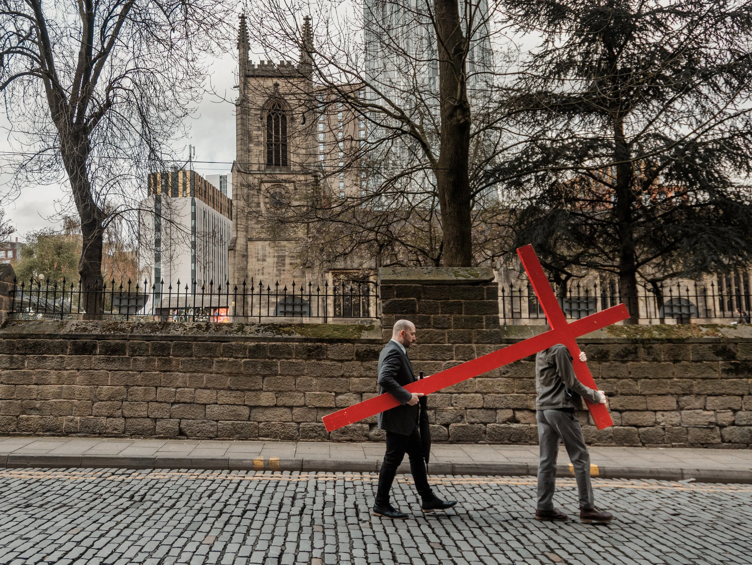 Two men walking along a cobblestone sidewalk, carrying a large red cross sign, with leafless trees and a stone church tower in the background. Theyre in front of st johns church in Leeds city centre