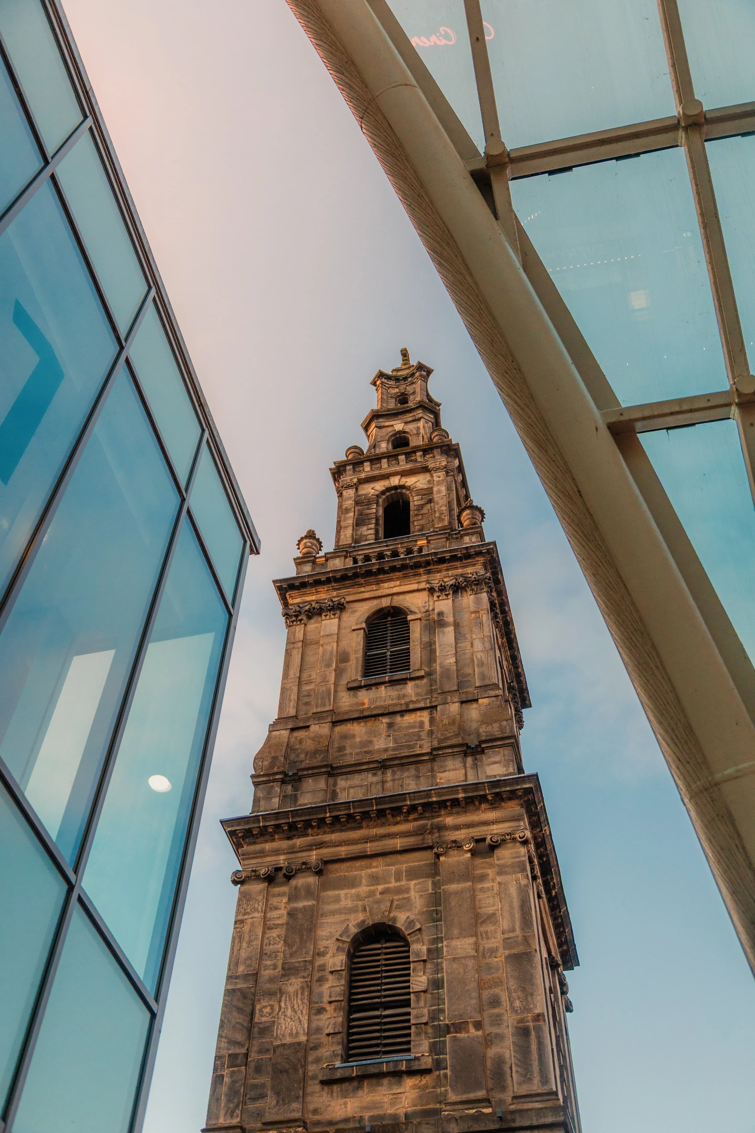 A tall historic clock tower viewed from ground level, flanked by modern glass buildings, under a partly cloudy sky. Leeds holy trinity church on boar lane
