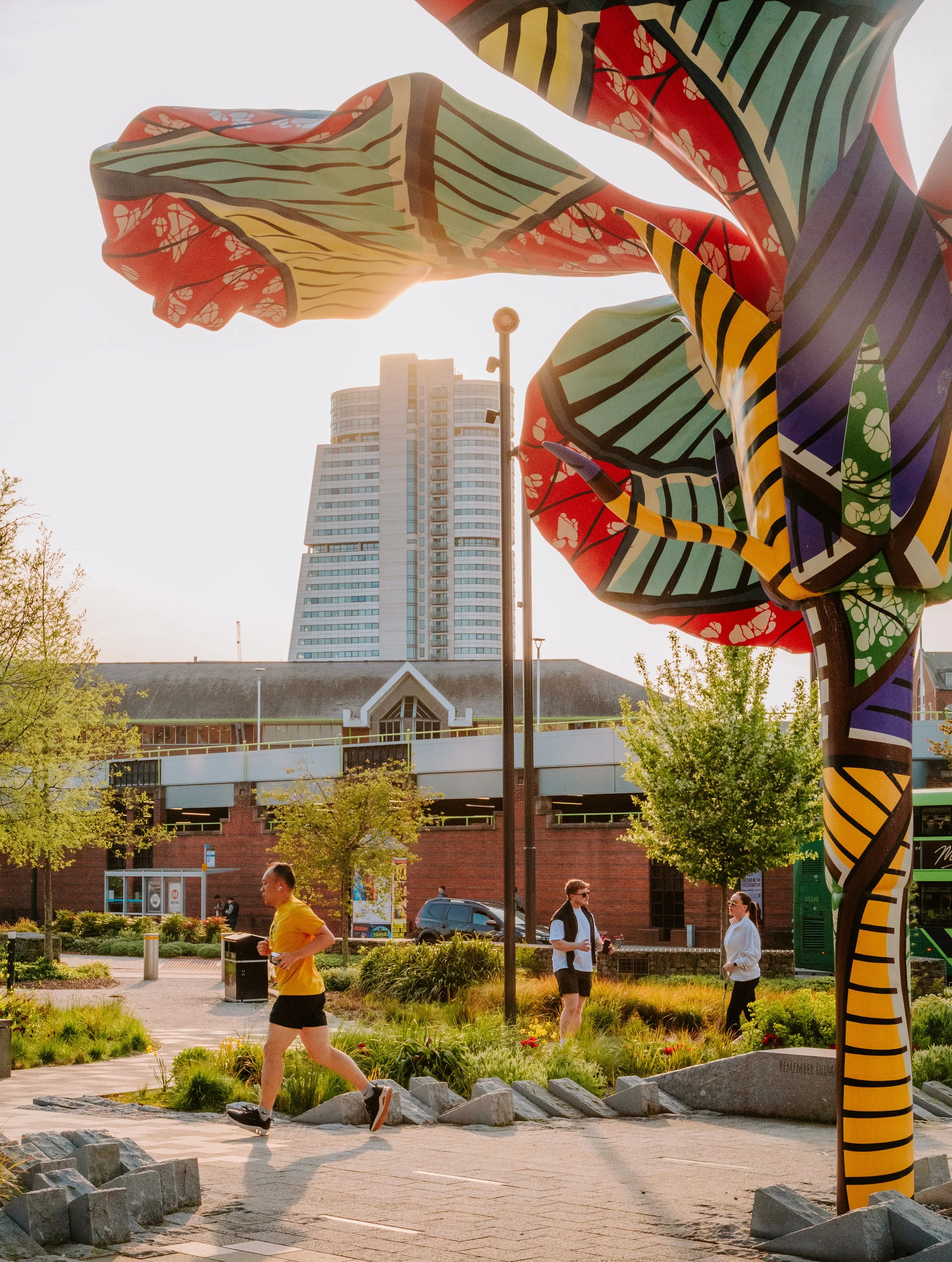 People jogging and walking in a park area with a large colorful abstract sculpture, trees, and a high-rise building in the background, under a clear sky with sunlight.