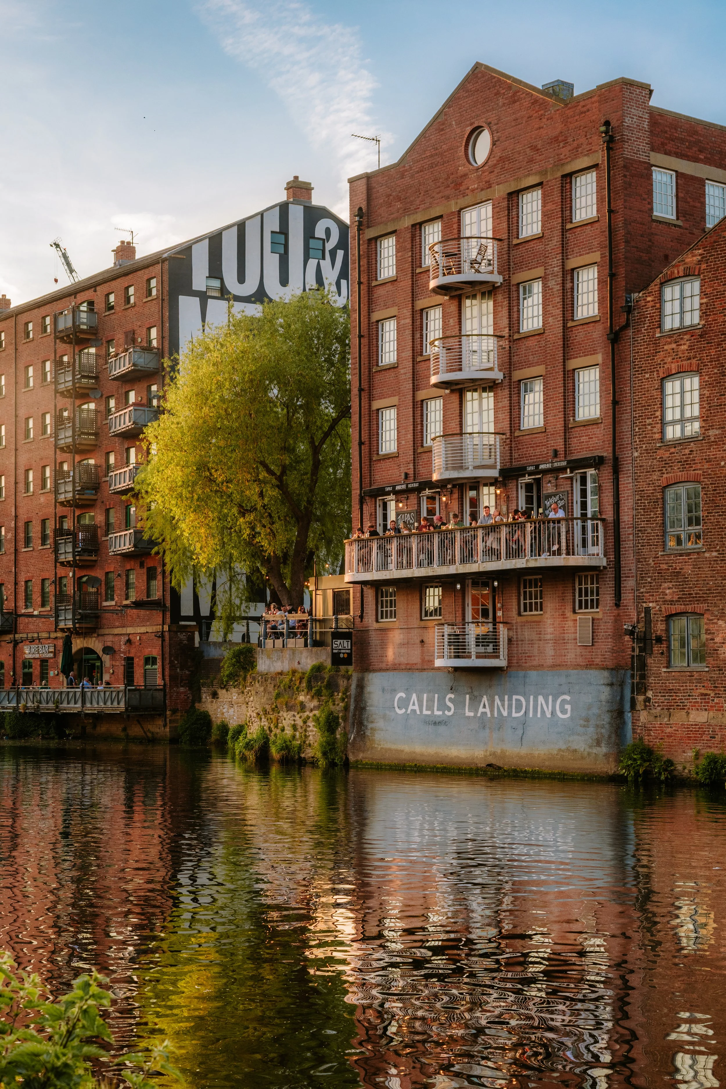Red brick apartment buildings along a river with balconies and a large mural featuring the letters "LOFT" painted on the side. The lower part of one building has the words "CALLS LANDING" . leeds city centre river aire canal sunset.