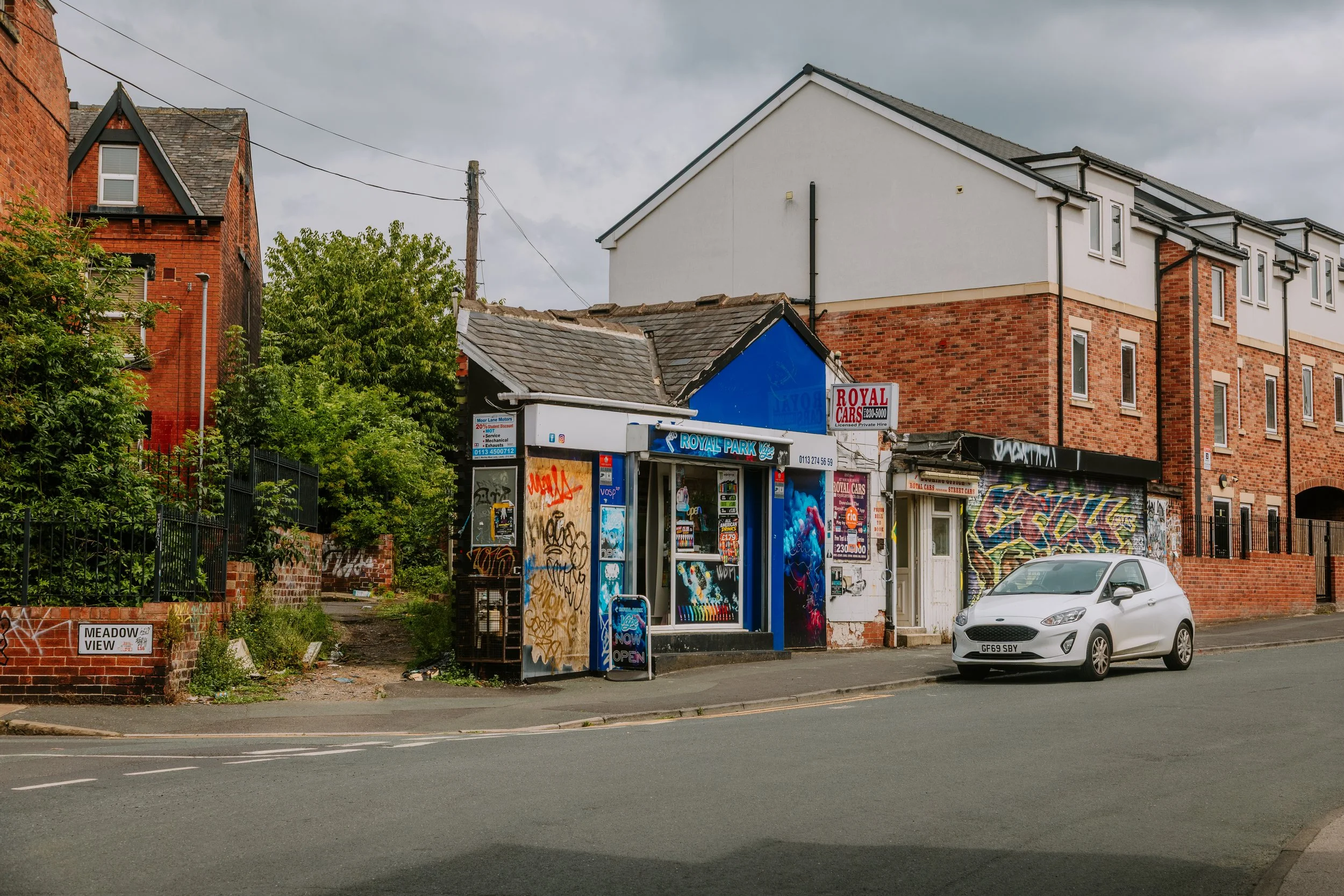 A street scene featuring a white car parked in front of a small shop with graffiti-covered walls. The shop has a sign for 'Royal Cars' and an 'Open' sign. Behind the shop are red brick residential buildings with multiple windows. Hyde park leeds 
