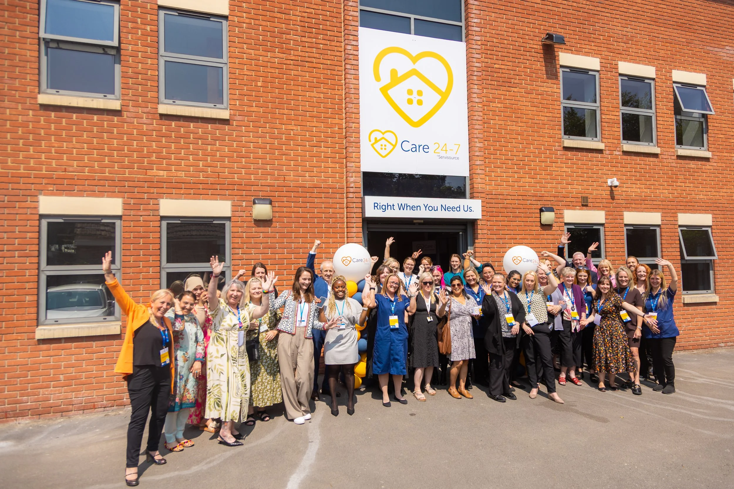 A large group of people gathered outside a brick building, celebrating. The building has multiple windows and a large sign showing a house icon with a heart and the words 'Care 24-7' and a slogan 'Right When You Need Us'. The crowd is smiling, waving