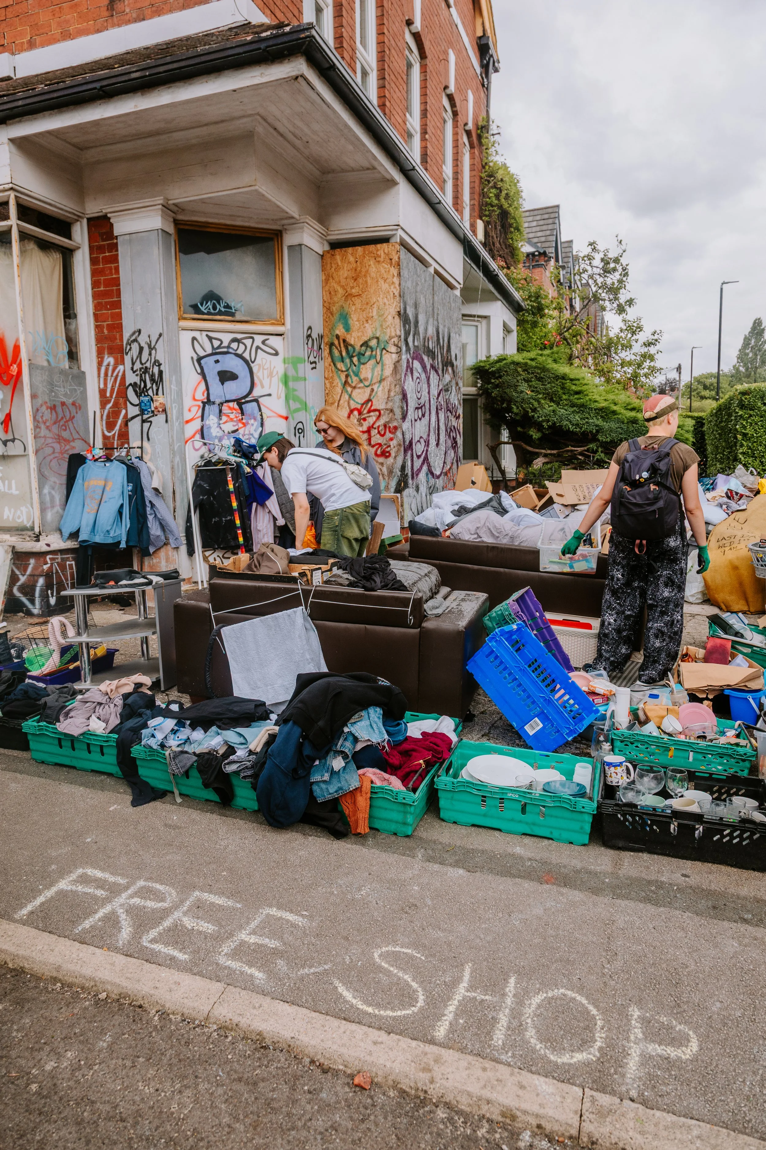 Street flea market with clothing, household items, and glassware on display next to a graffiti-covered building. Hyde park free shop.
