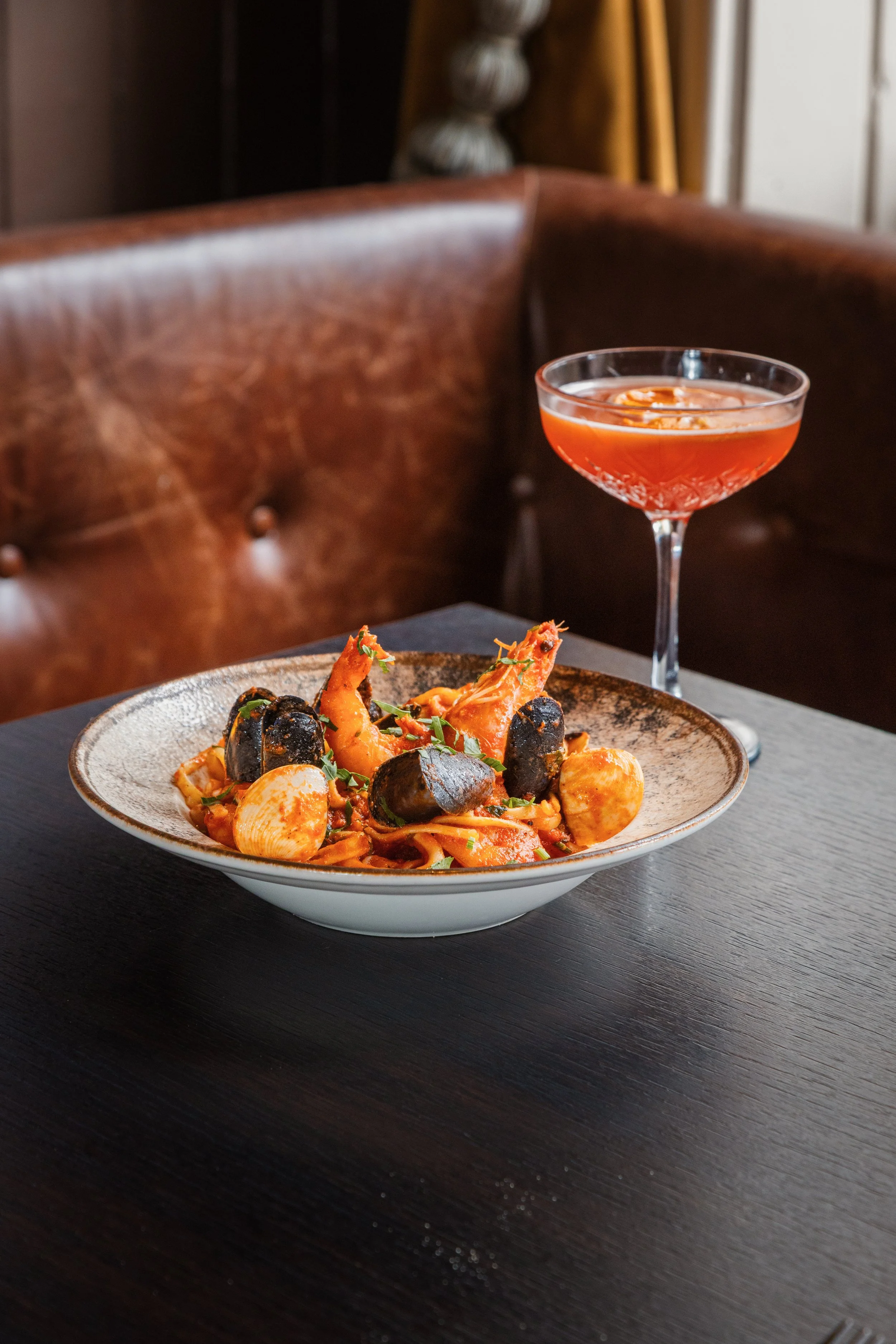 A bowl of seafood pasta with shrimp, mussels, and clams, garnished with herbs, on a dark wooden table. In the background, a cocktail glass filled with an orange-colored drink is visible. york leeds yorkshire restaurant photographer videographer